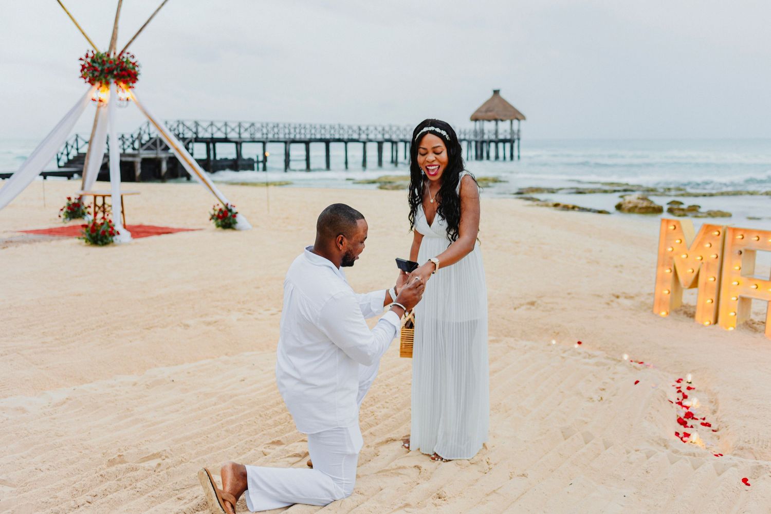 Beach proposal scene with white decor and rose petals on sandy shore.