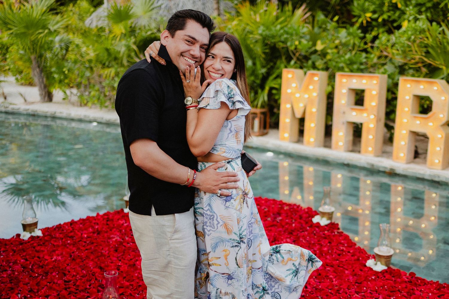 Couple embracing by a pool surrounded by red rose petals at sunset.