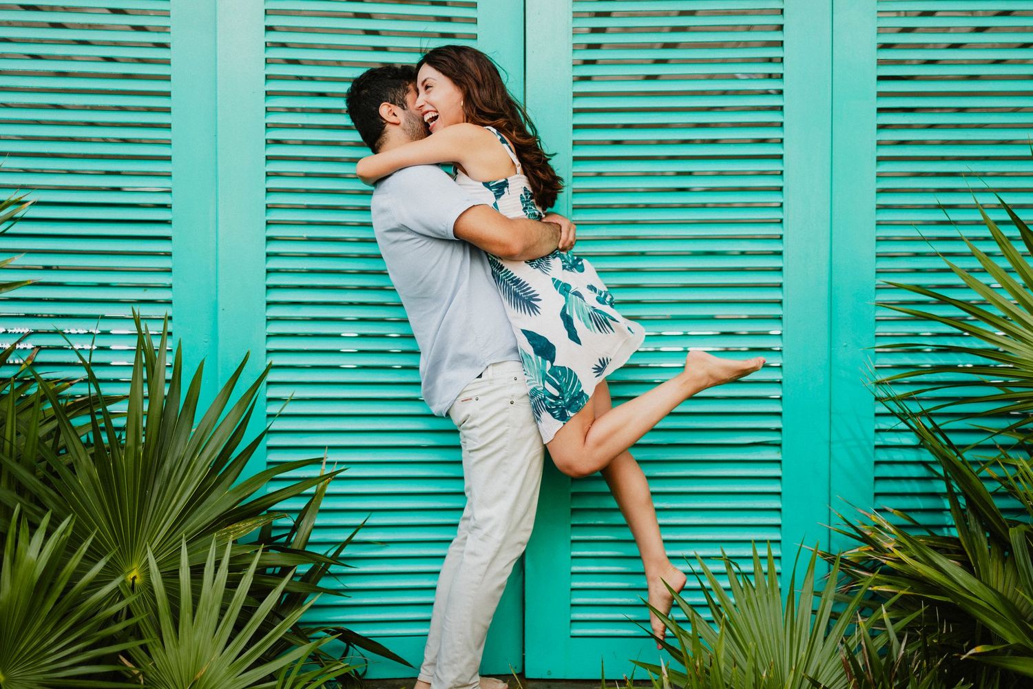 Romantic couple embracing against turquoise shutters with tropical plants.