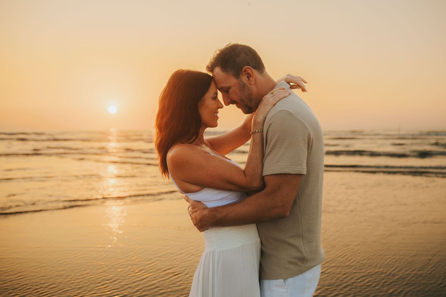 Two people embrace on a sandy beach at sunset while gentle waves roll onto the shore in the background.