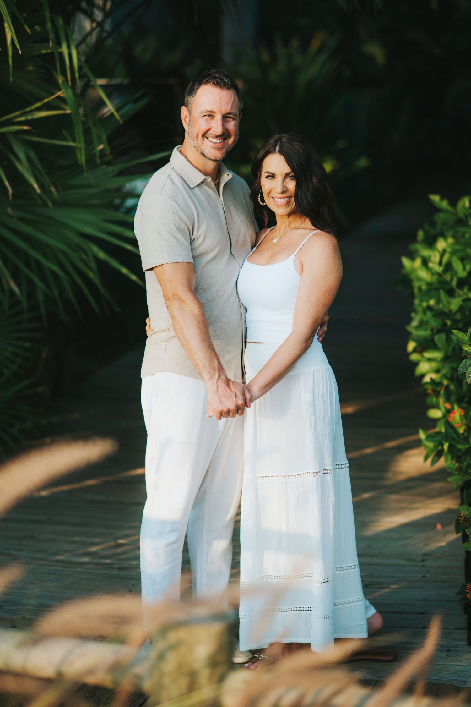 Couple in white outfits posing together in a lush tropical garden setting.