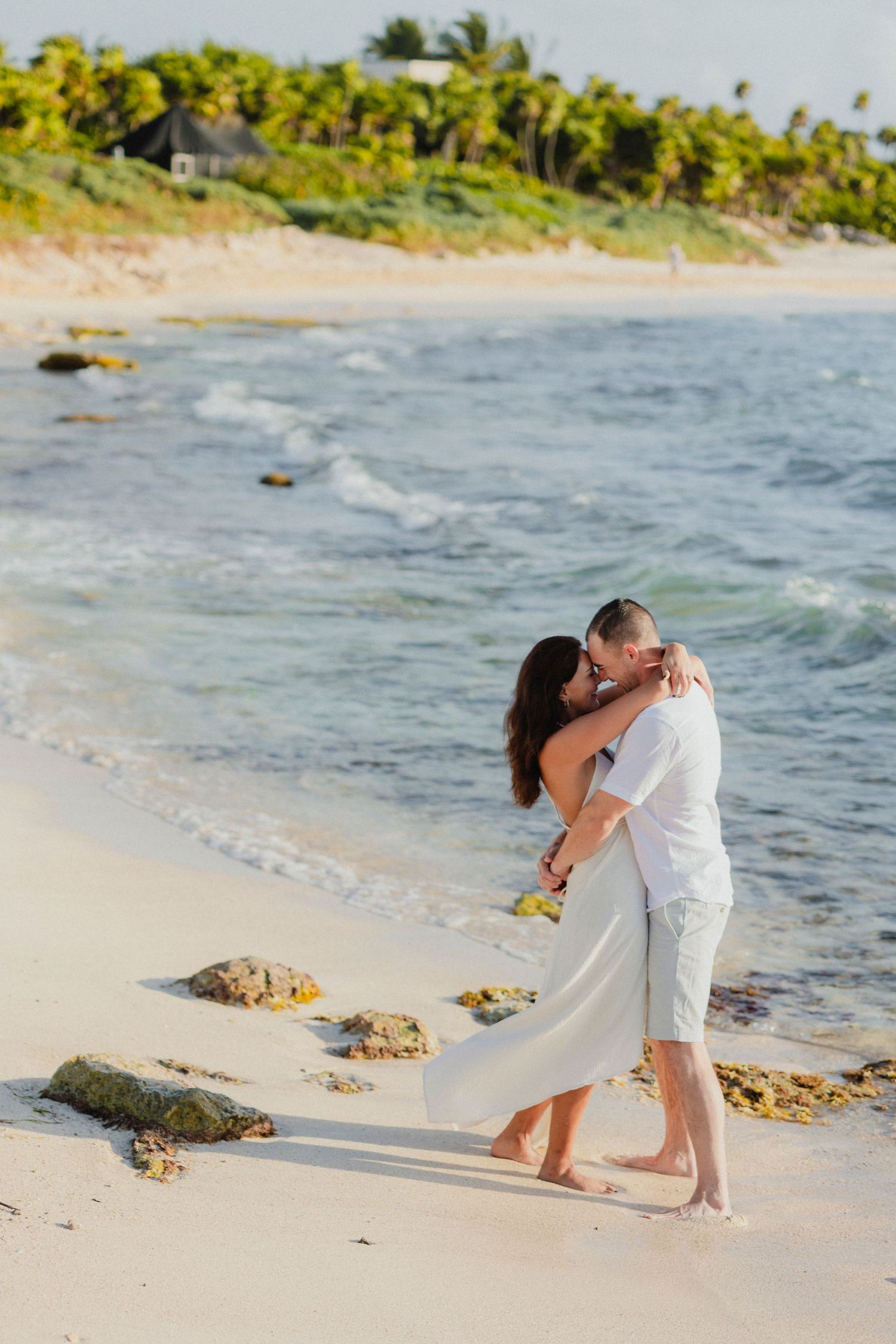 Couple in white embracing on tropical beach with waves and palm trees.