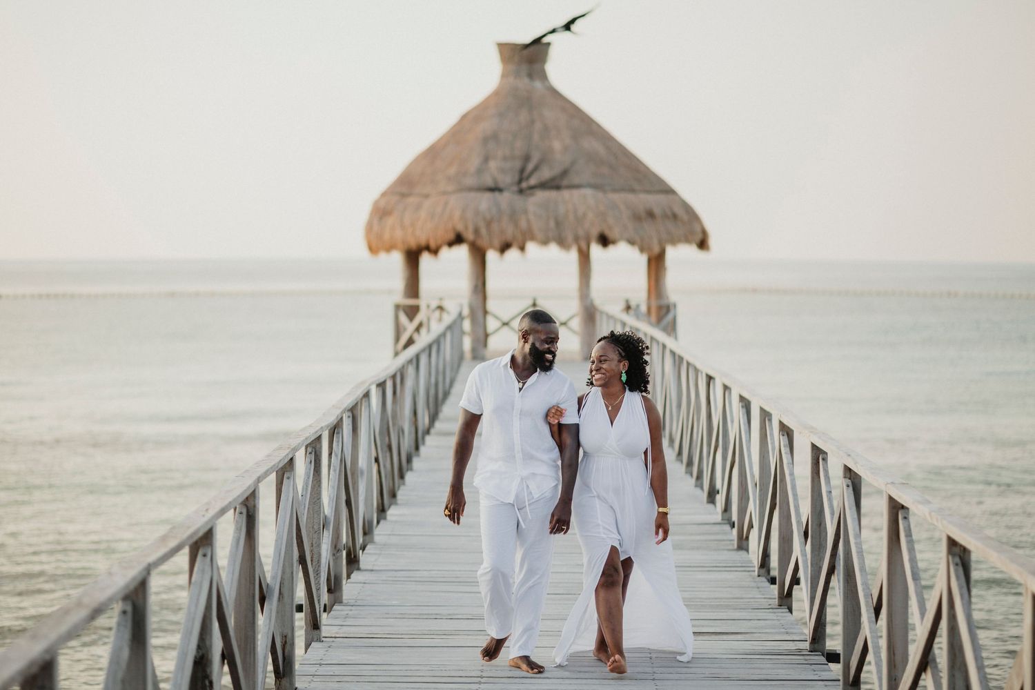Wedding party walking along wooden pier with thatched roof gazebo.
