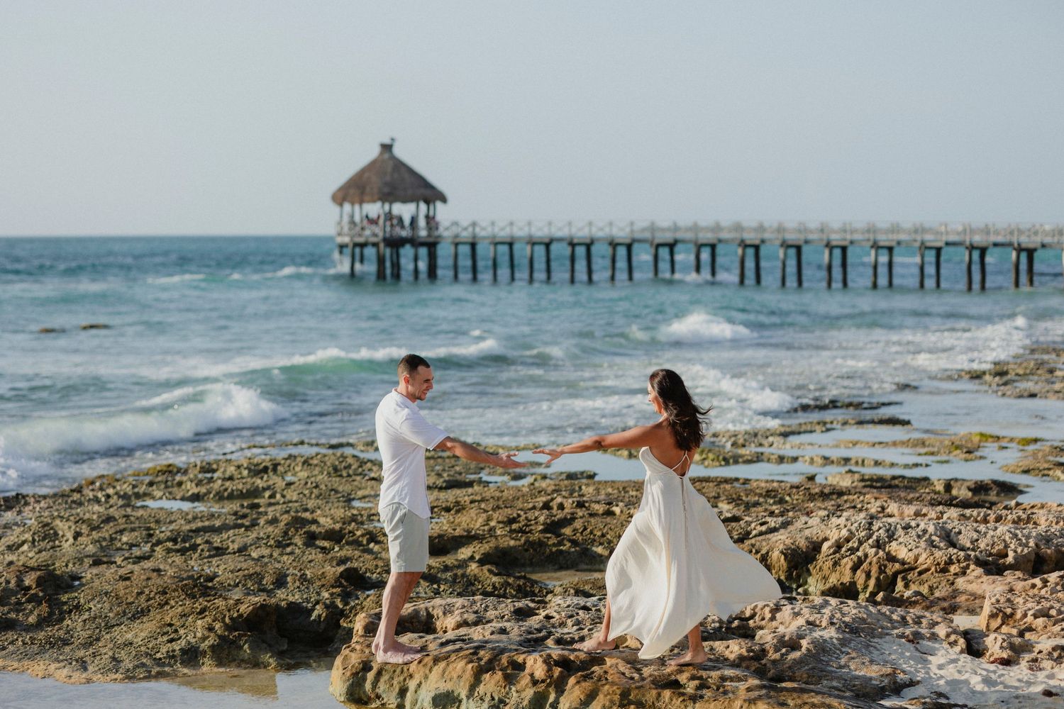Couple holding hands on rocky beach with pier and ocean waves in background.