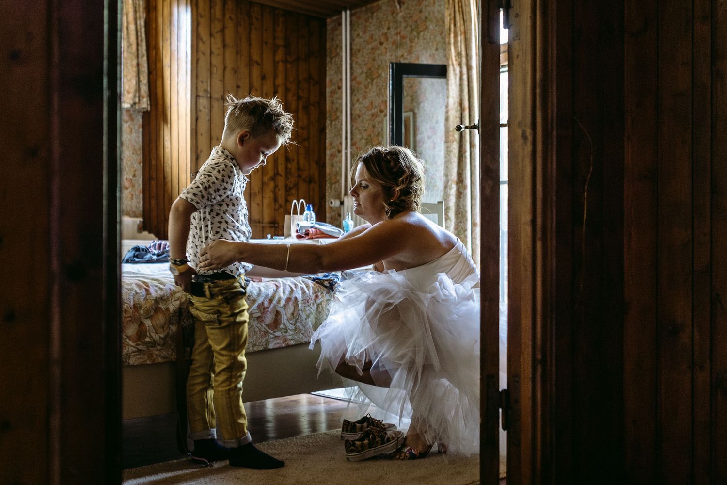 A bride in a flowing white dress stands elegantly in front of a window with natural light streaming in.