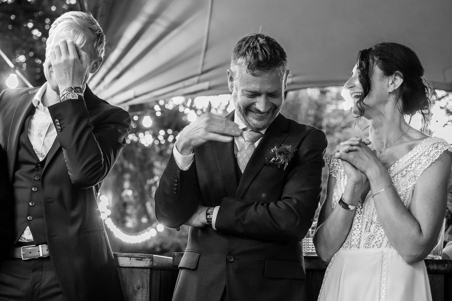 A candid black and white wedding reception moment showing joyful laughter between the couple and a guest.