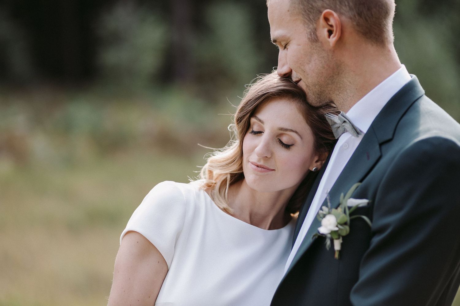 A romantic wedding couple shares an intimate moment outdoors surrounded by greenery and natural light.
