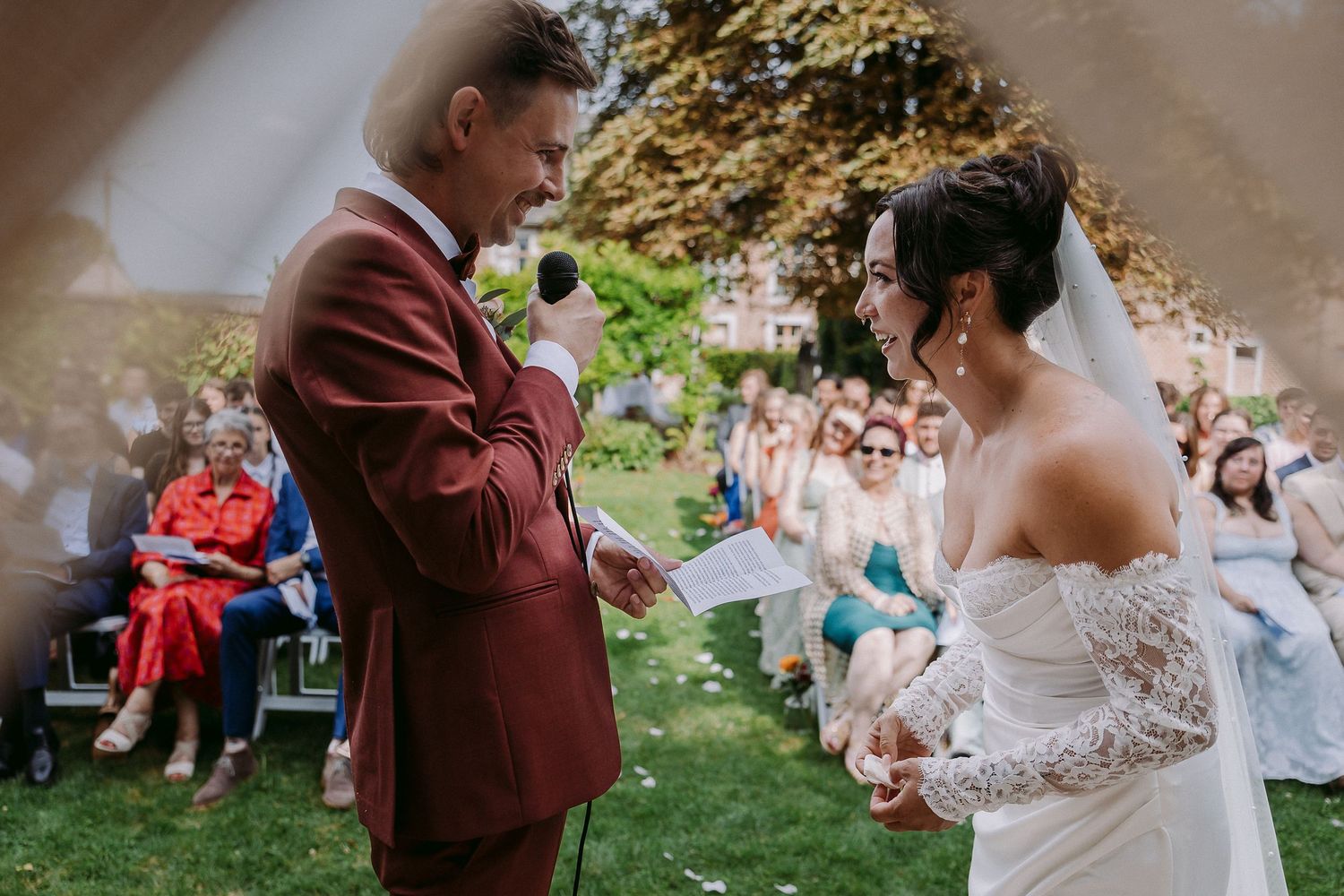 Wedding guests sit on blankets and chairs during an outdoor garden ceremony while couple exchanges vows.