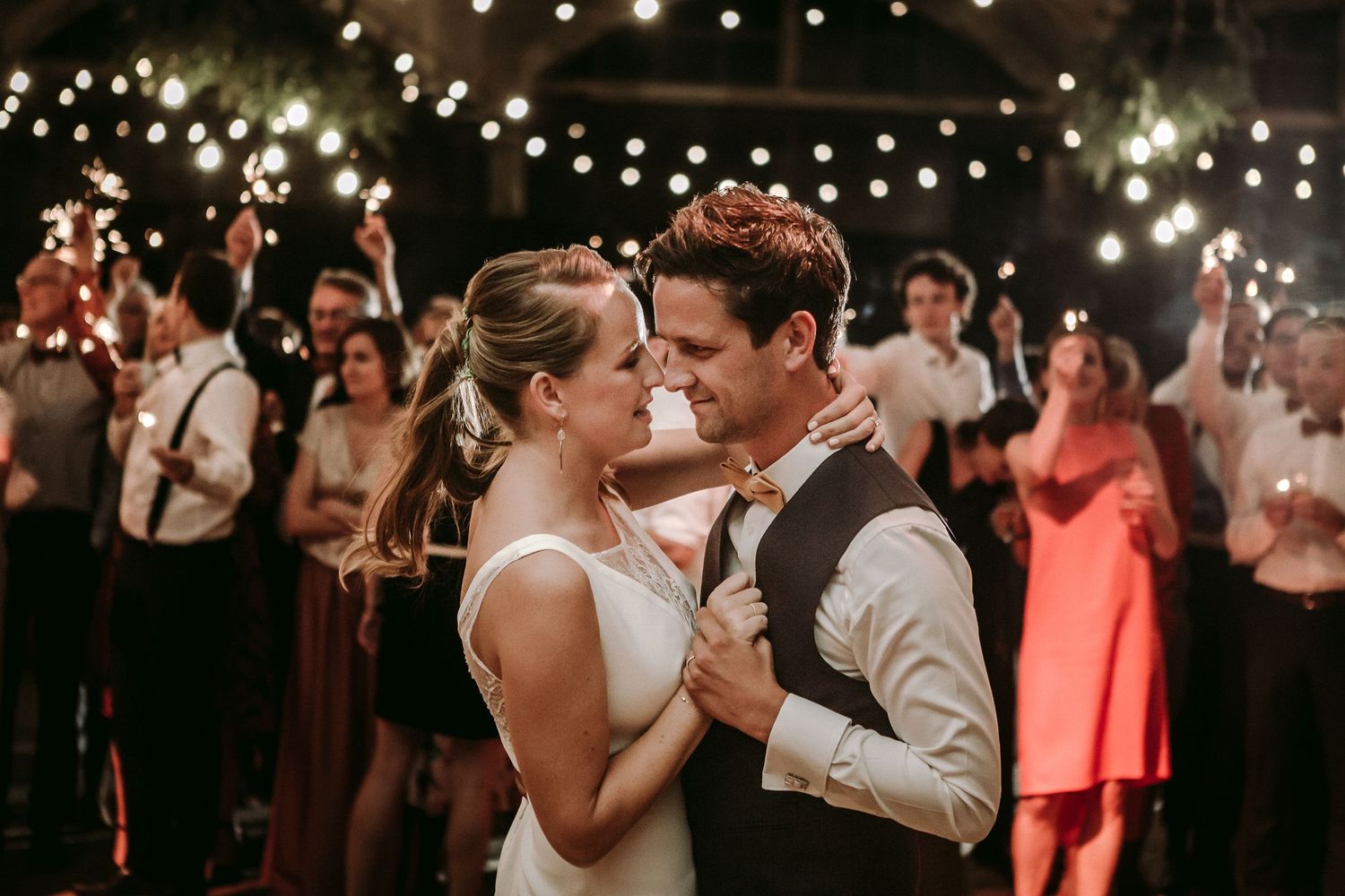 A couple shares an intimate dance moment at a wedding reception under twinkling string lights with guests in the background.
