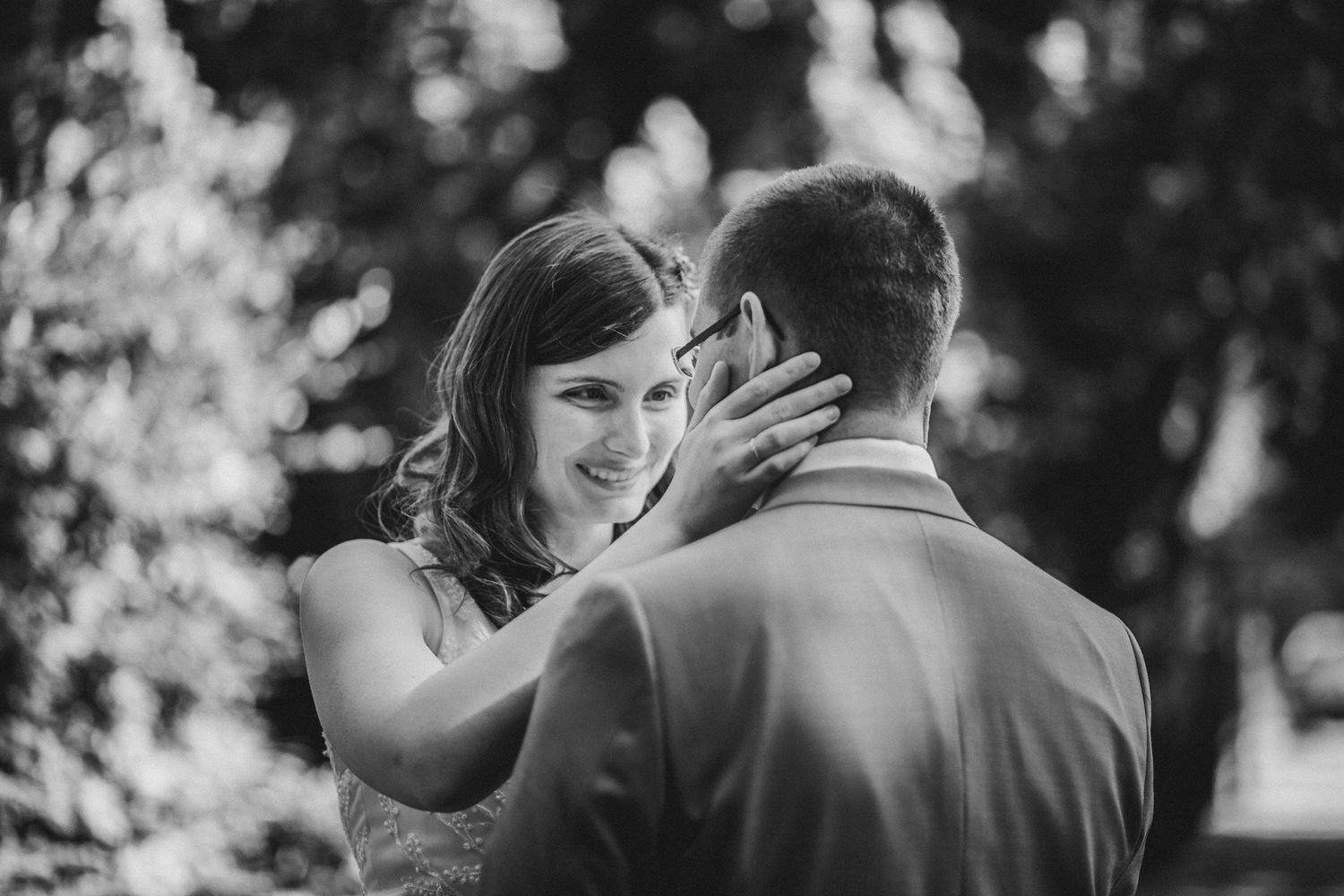 A tender black and white moment between a couple embracing outdoors with sunlit trees in the background.
