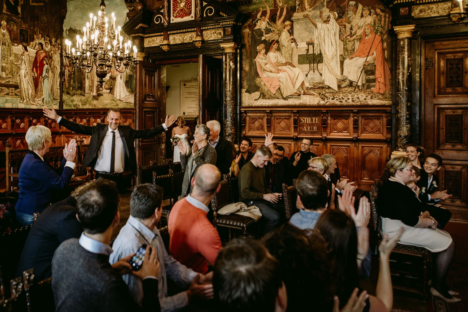 Patrons gather at a historic bar with ornate decor, vintage chandeliers and dark wood paneling.