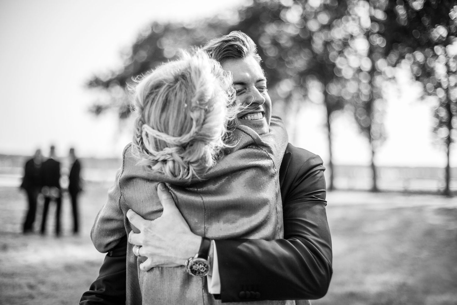 Black and white photo showing an intimate embrace between two people outdoors with blurred figures in background.