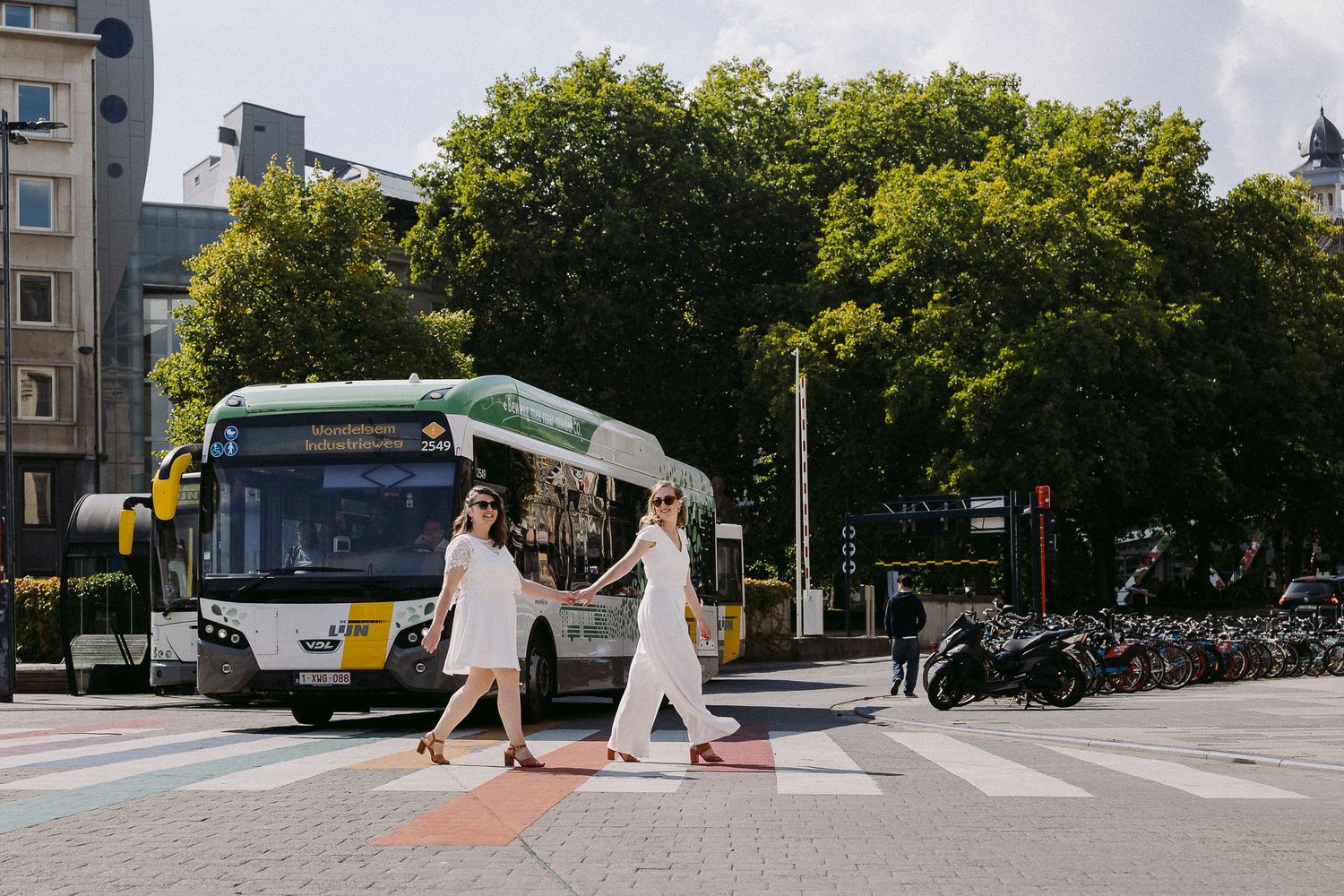 People in white dresses walk across a crosswalk in an urban setting with trees and buildings in the background.