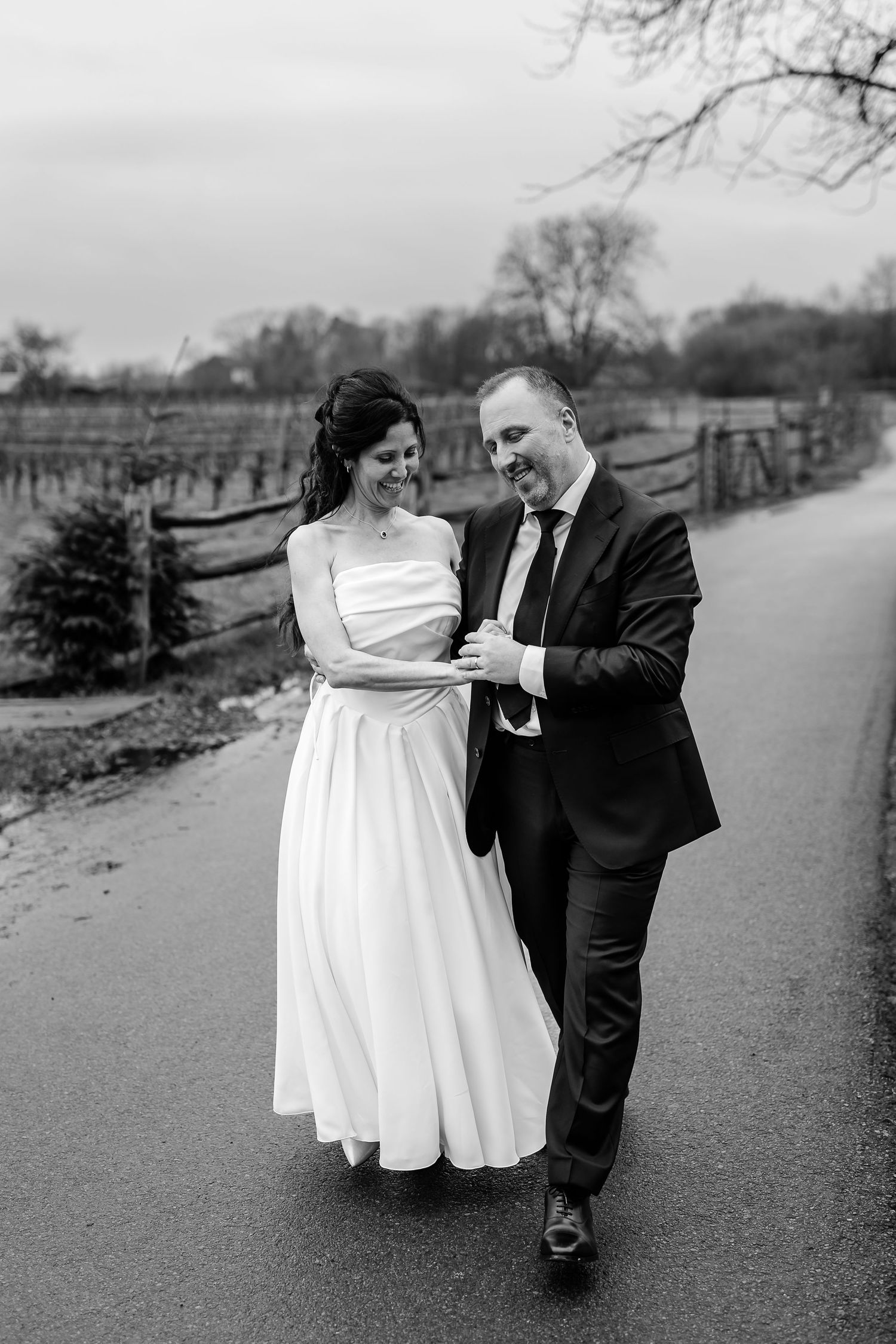 A black and white wedding photo of a couple walking arm in arm down a path with bare trees in the background.