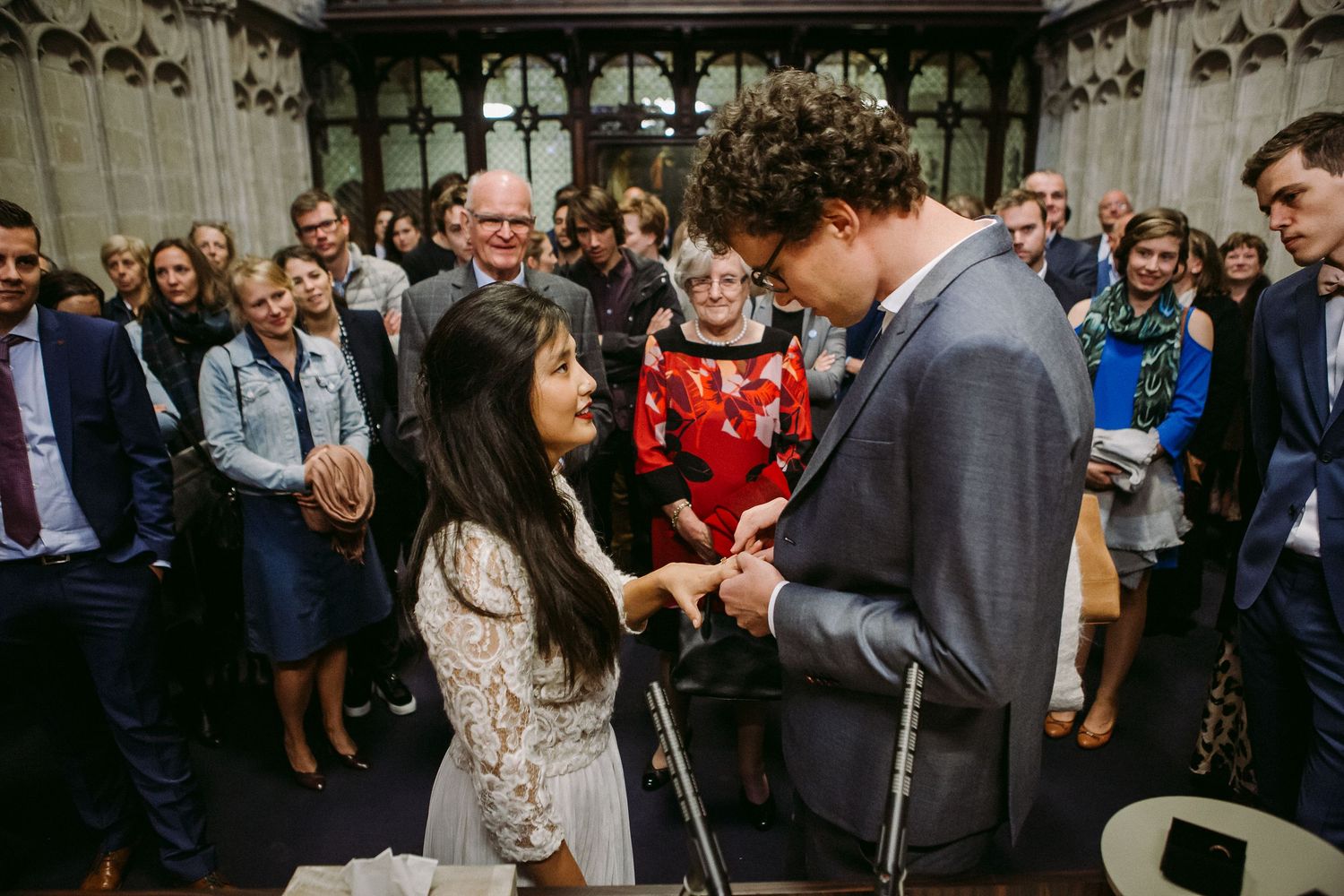 A couple exchanges rings during an intimate wedding ceremony in a historic chapel surrounded by guests.