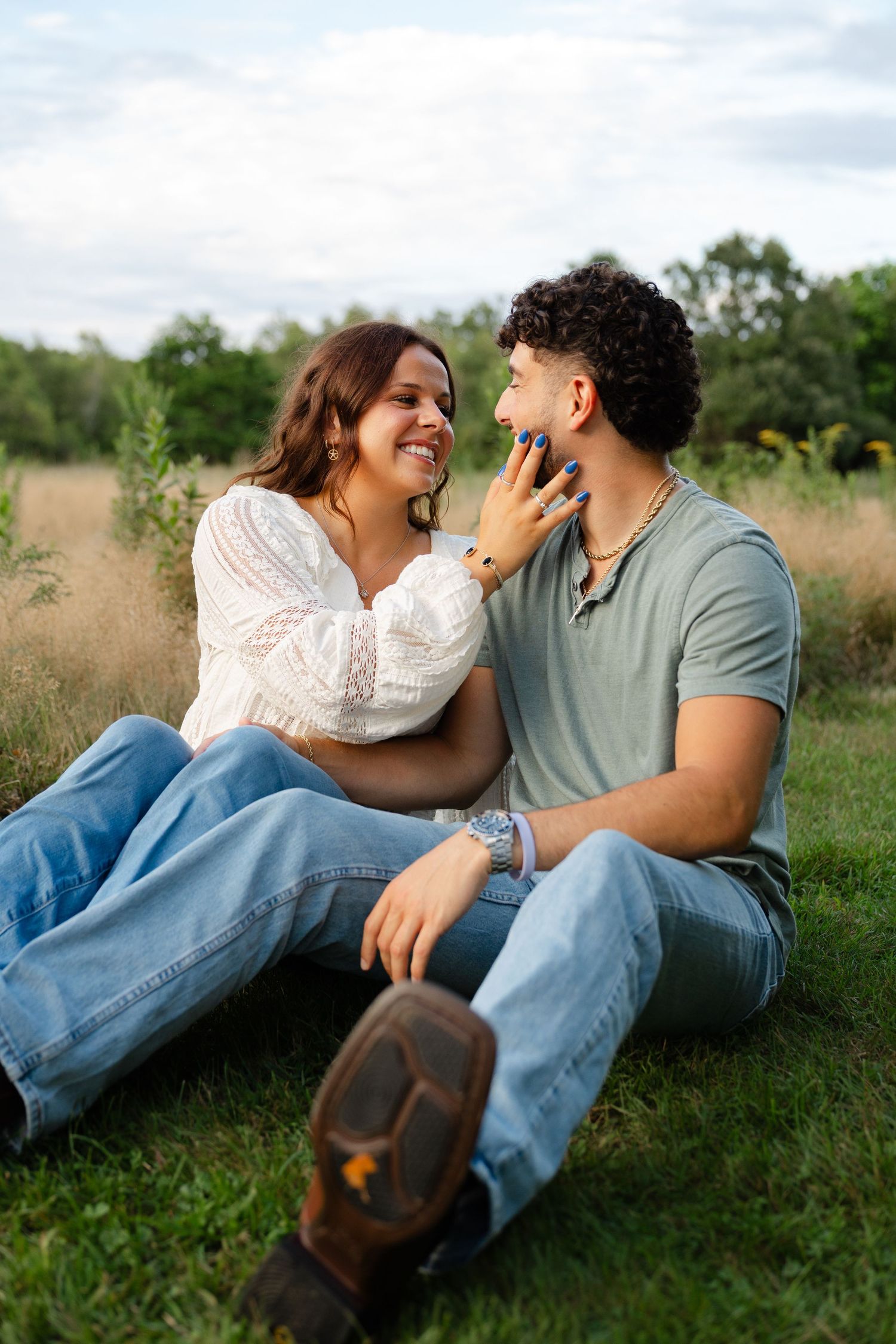 A couple sitting on grass, smiling at each other, woman in white top touching man's face, trees in background.
