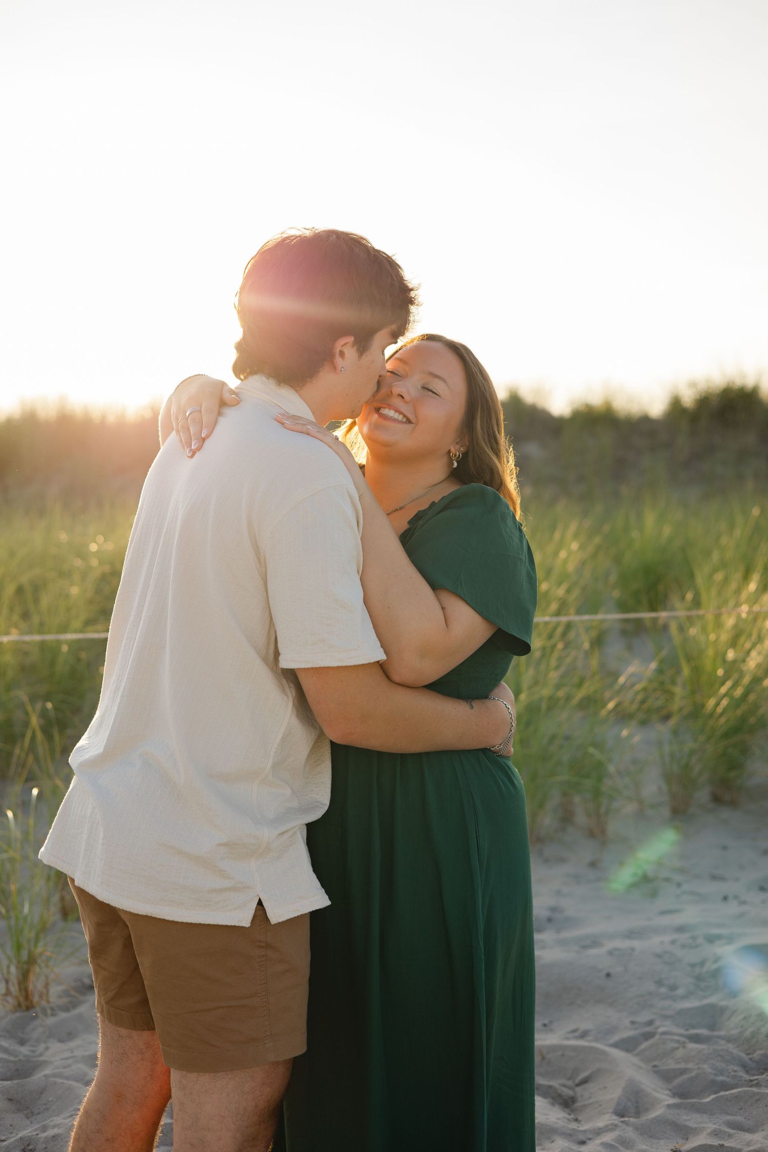 Couple embracing at sunset on a sandy beach, woman in green dress smiling joyfully, golden backlight glow.