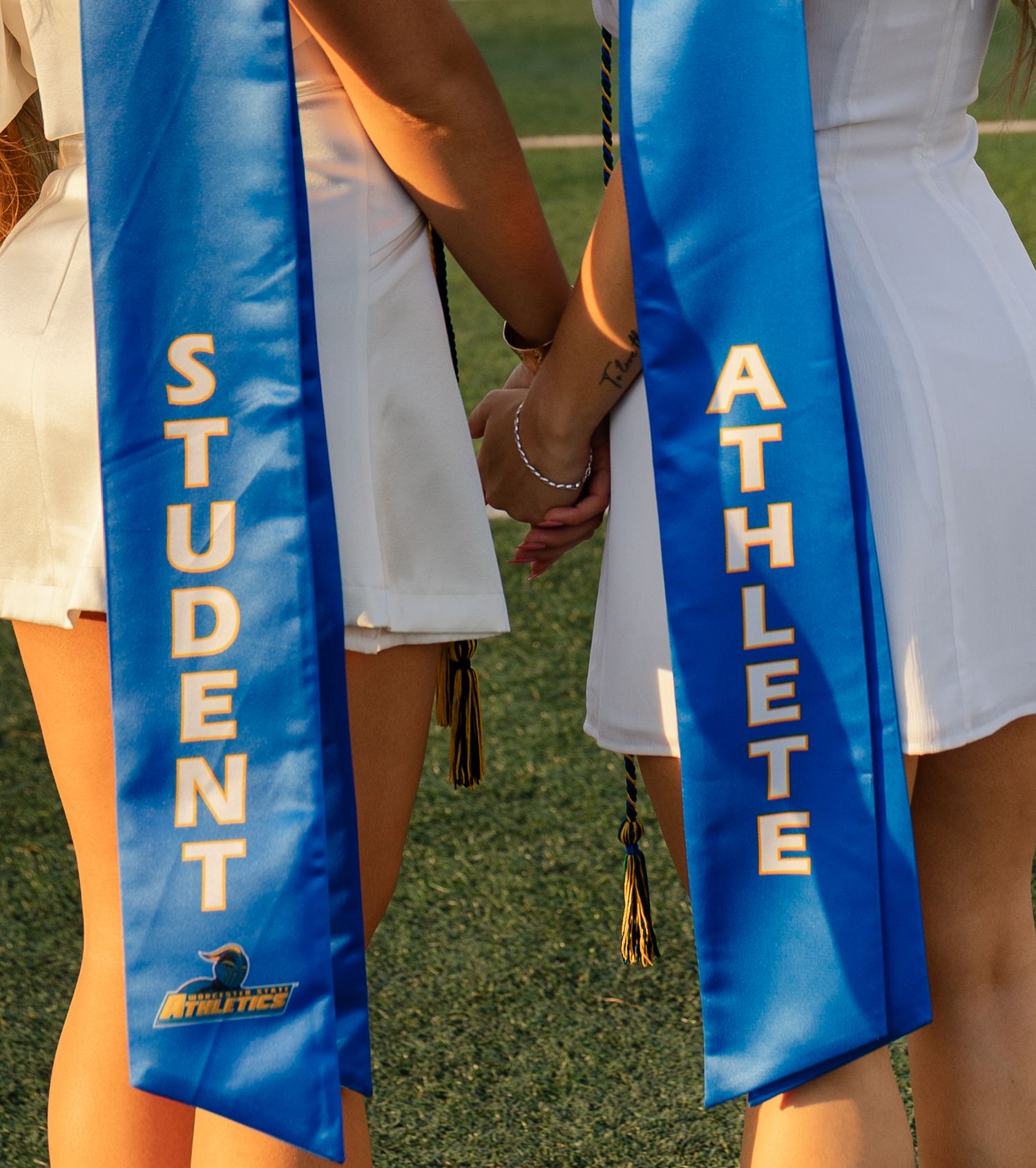 Two graduates wearing blue Student Athlete sashes hold hands on a field during a graduation ceremony.