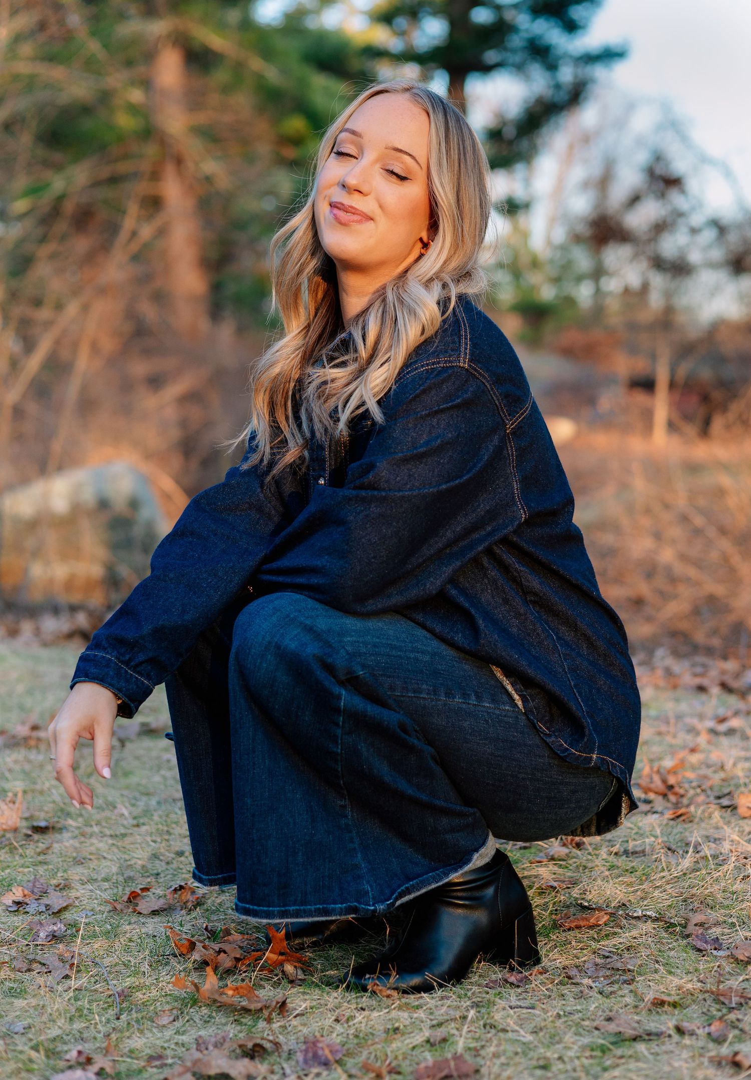 Woman in dark denim jacket and jeans crouching outdoors in a field during golden hour.