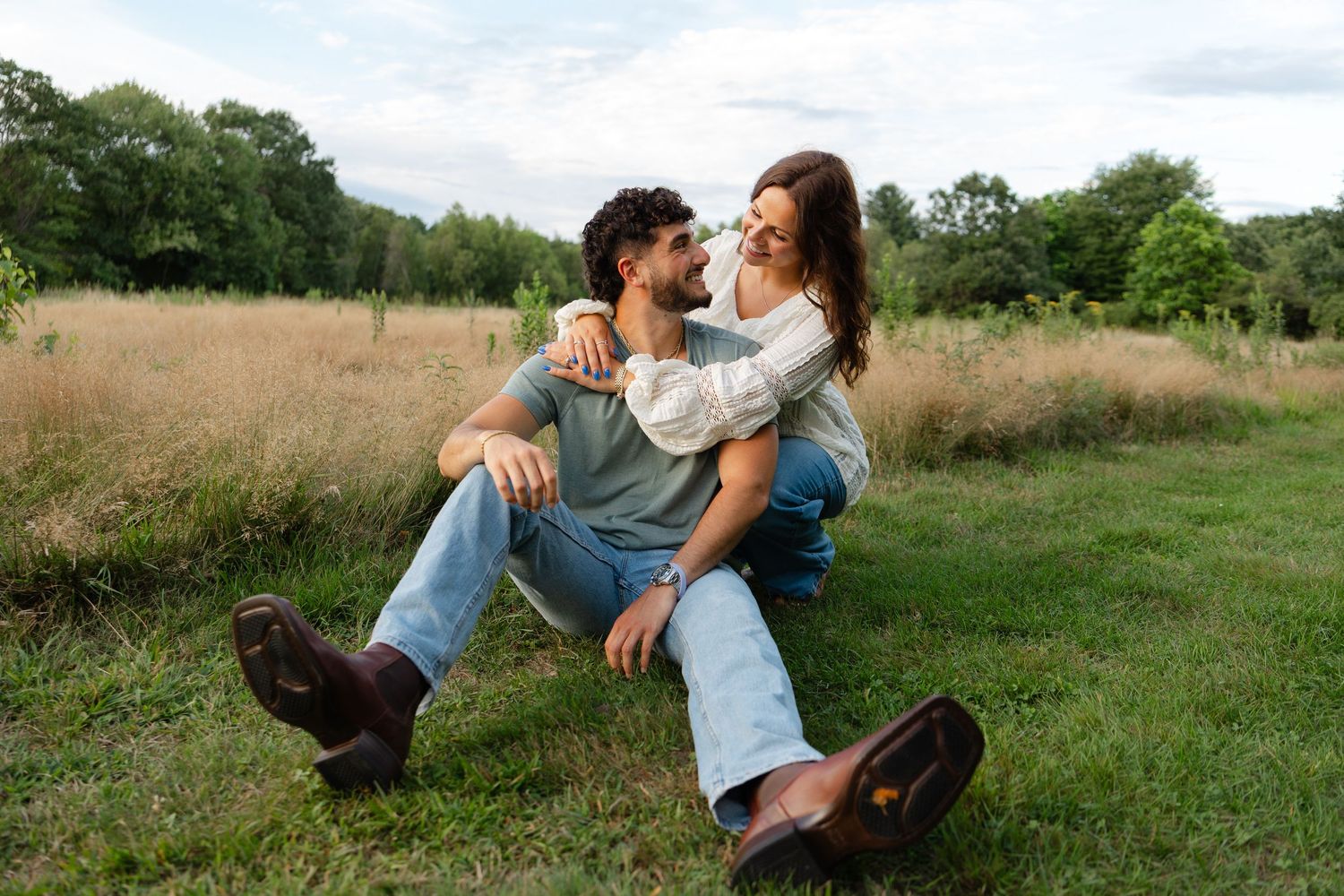 Couple sitting together on green grass in a field, sharing a romantic moment with trees in the background.