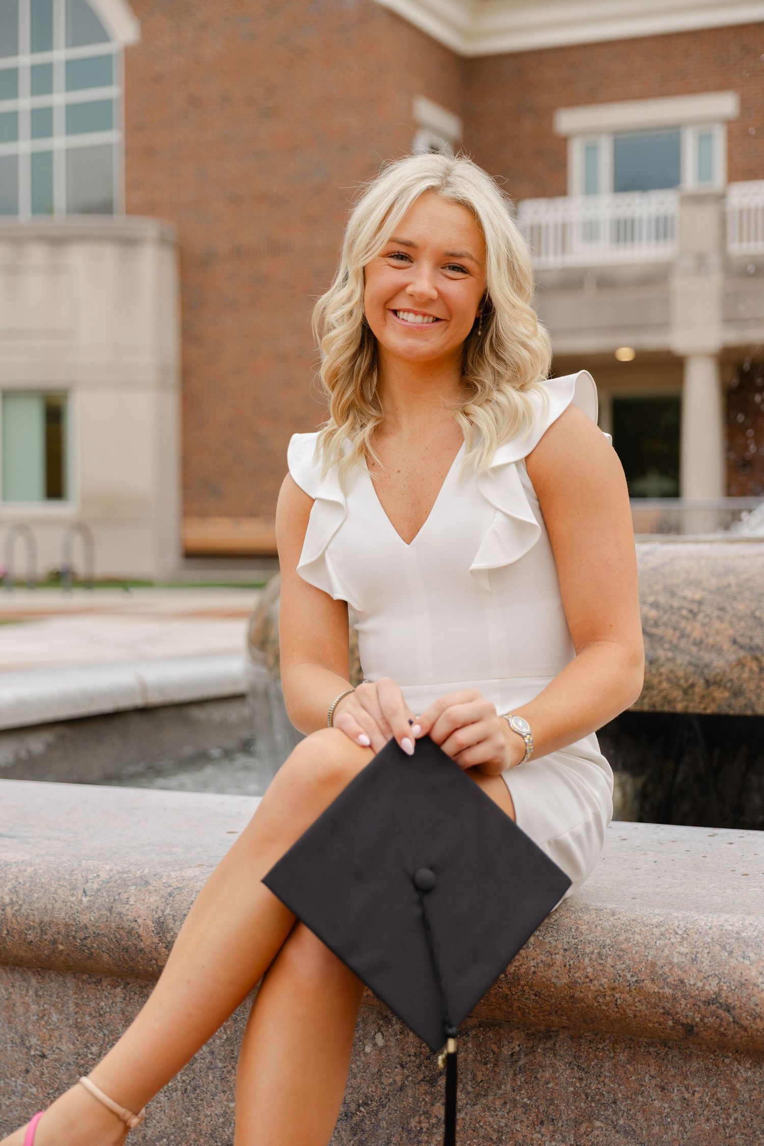 Smiling blonde graduate in white dress holding black mortarboard cap seated by campus fountain.