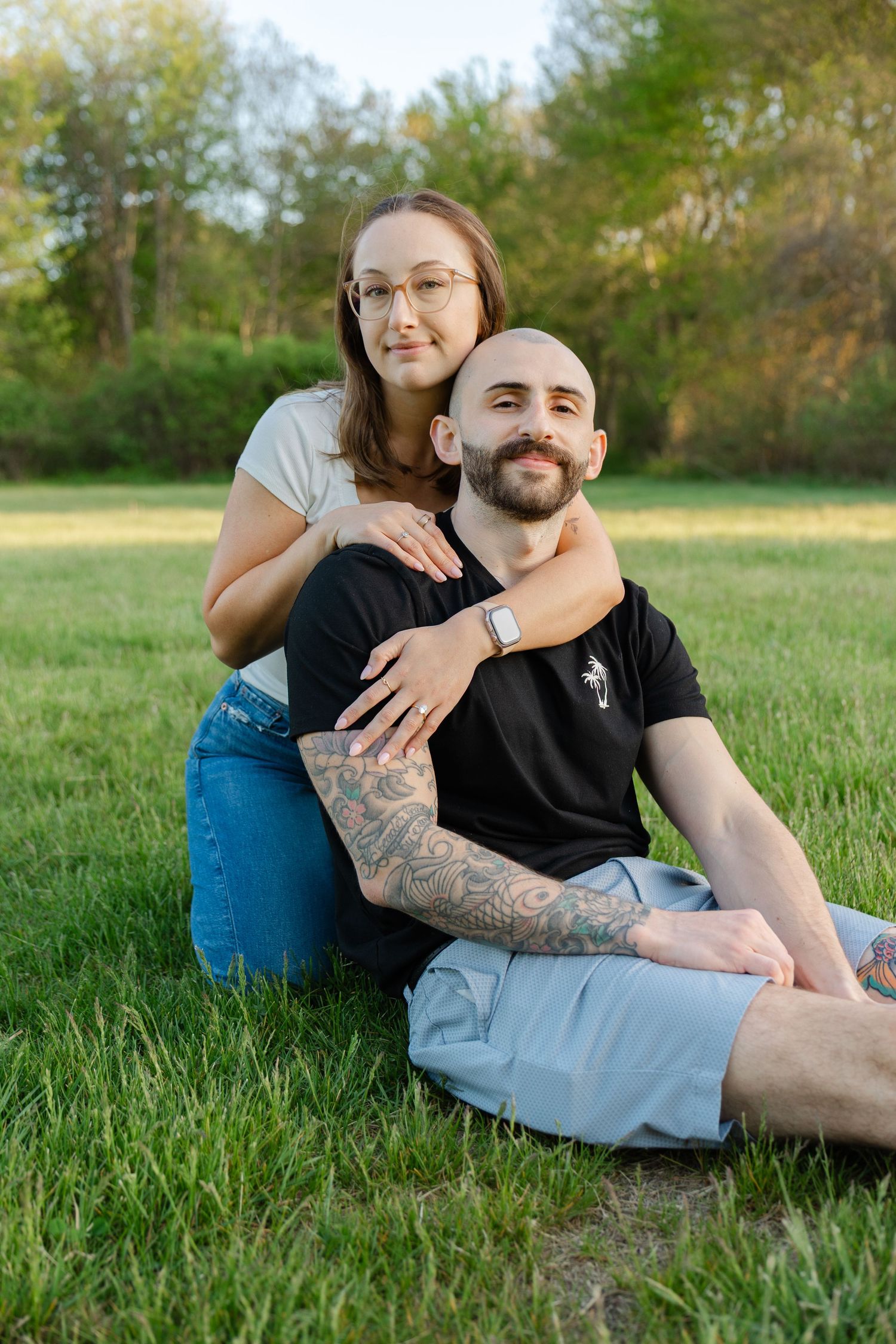 Happy couple sitting on green grass in park, woman with glasses embracing tattooed bald man, sunny day.
