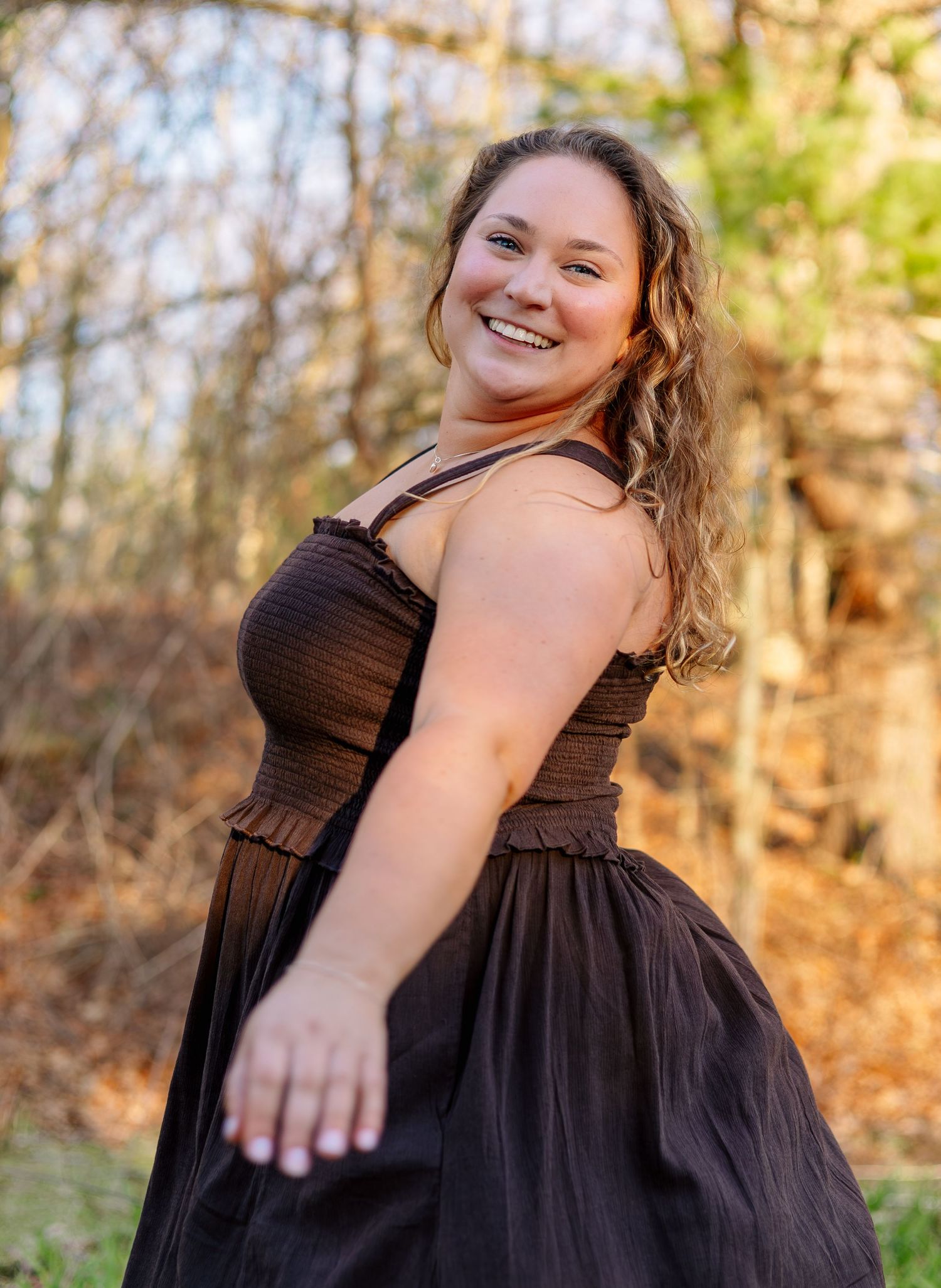 Smiling curly-haired woman in brown sundress twirling outdoors in wooded autumn setting.