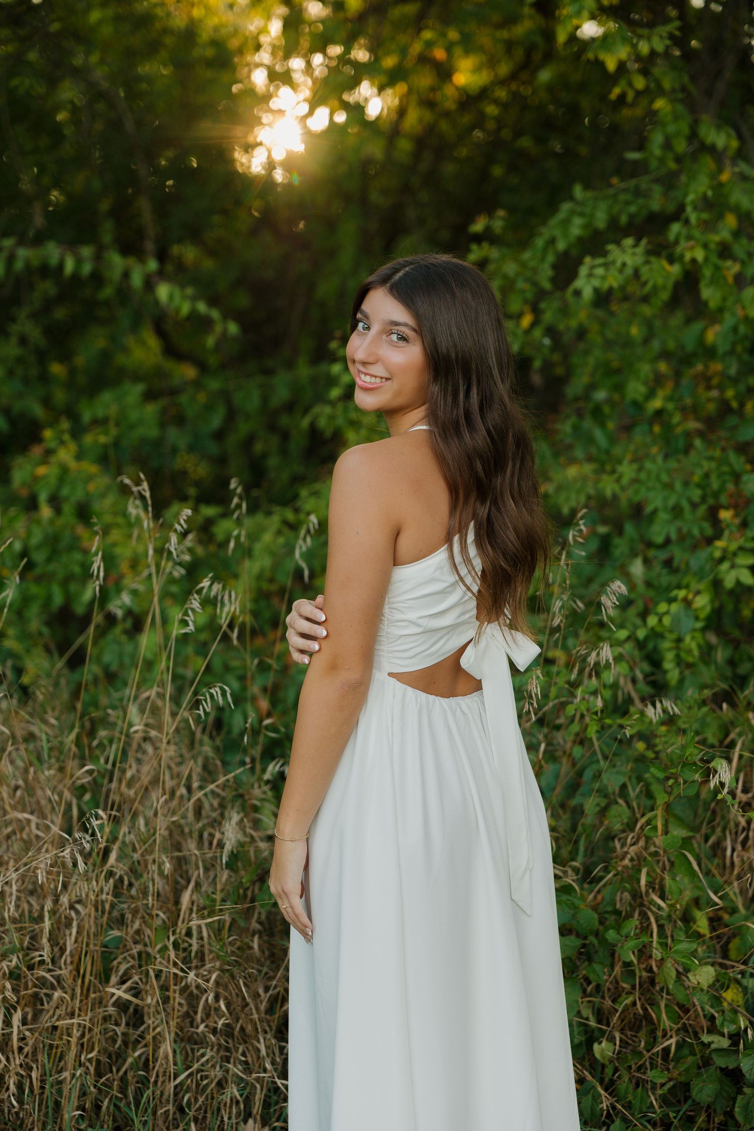 Young woman in white halter dress smiling over shoulder at golden hour sunset in lush green nature setting.