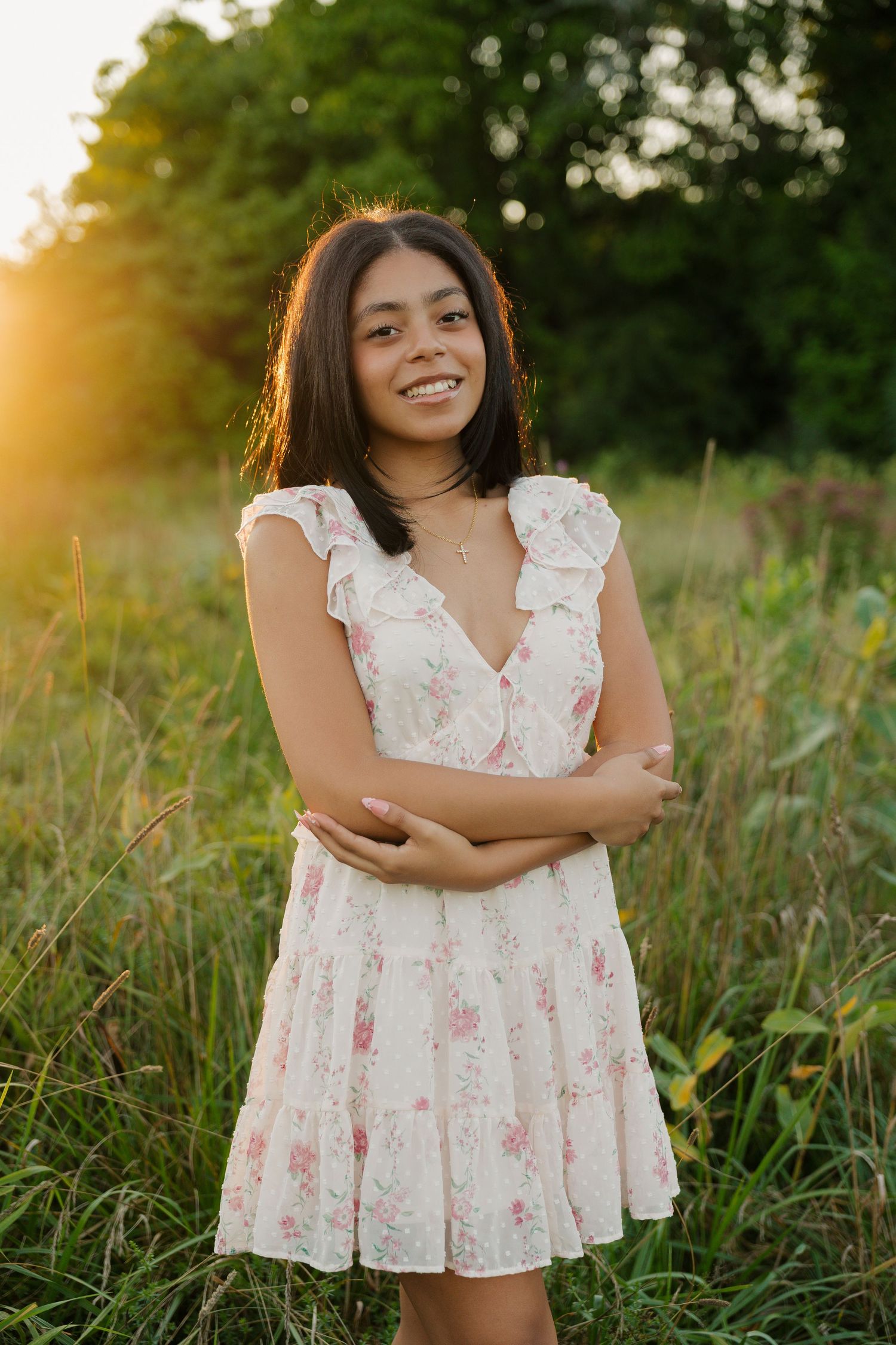 Young woman in floral white dress smiling in a sunlit meadow at golden hour.