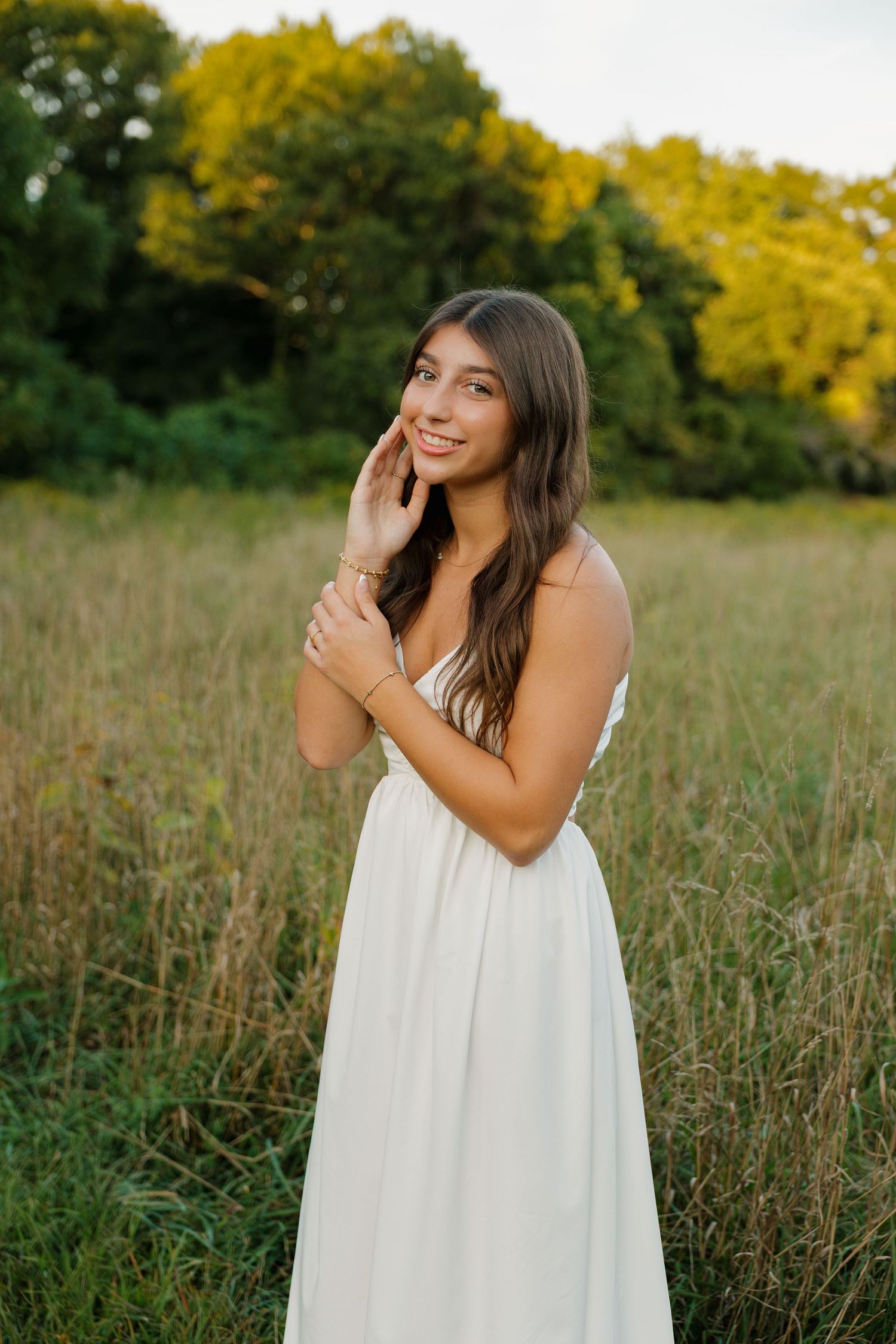 Young woman in white sundress posing in golden meadow with green trees, smiling softly at sunset.
