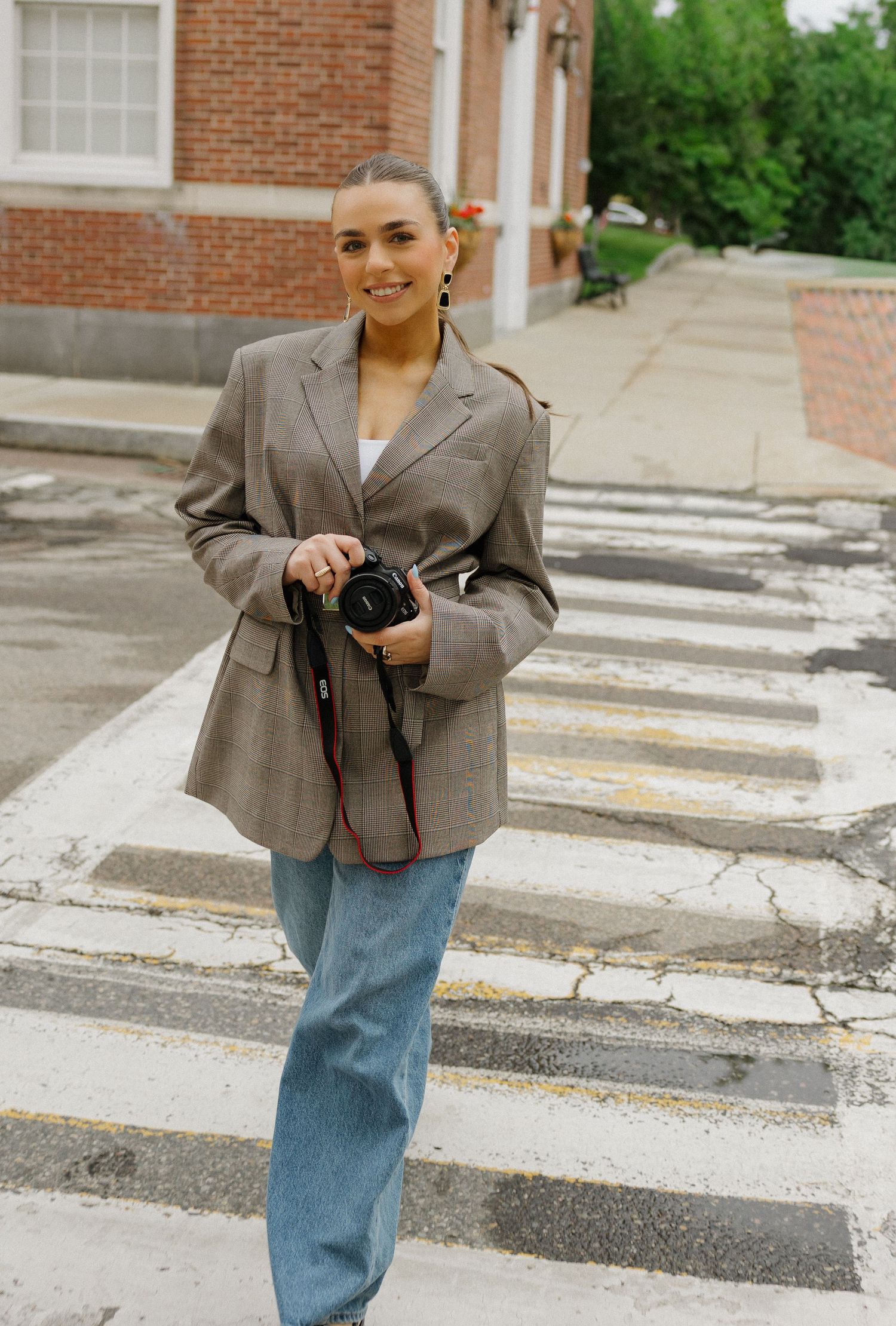 Woman in blazer and jeans holding a camera, smiling on a crosswalk in an urban setting.