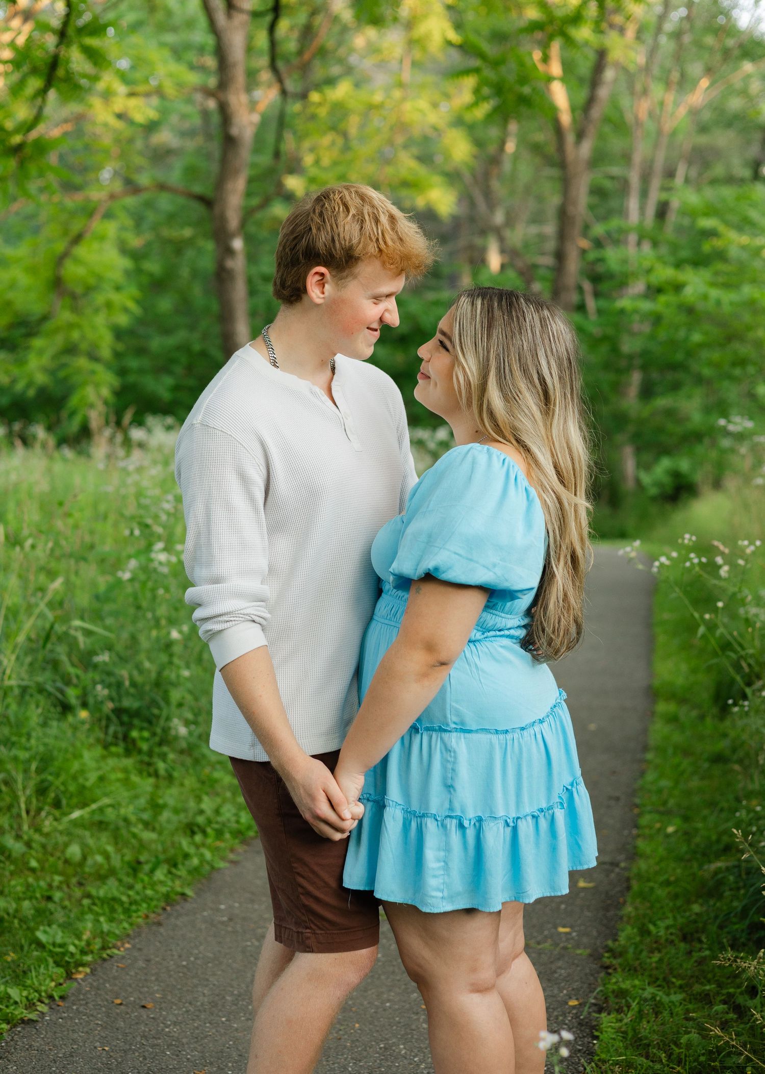 A young couple holds hands and gazes at each other on a scenic path surrounded by lush green trees.