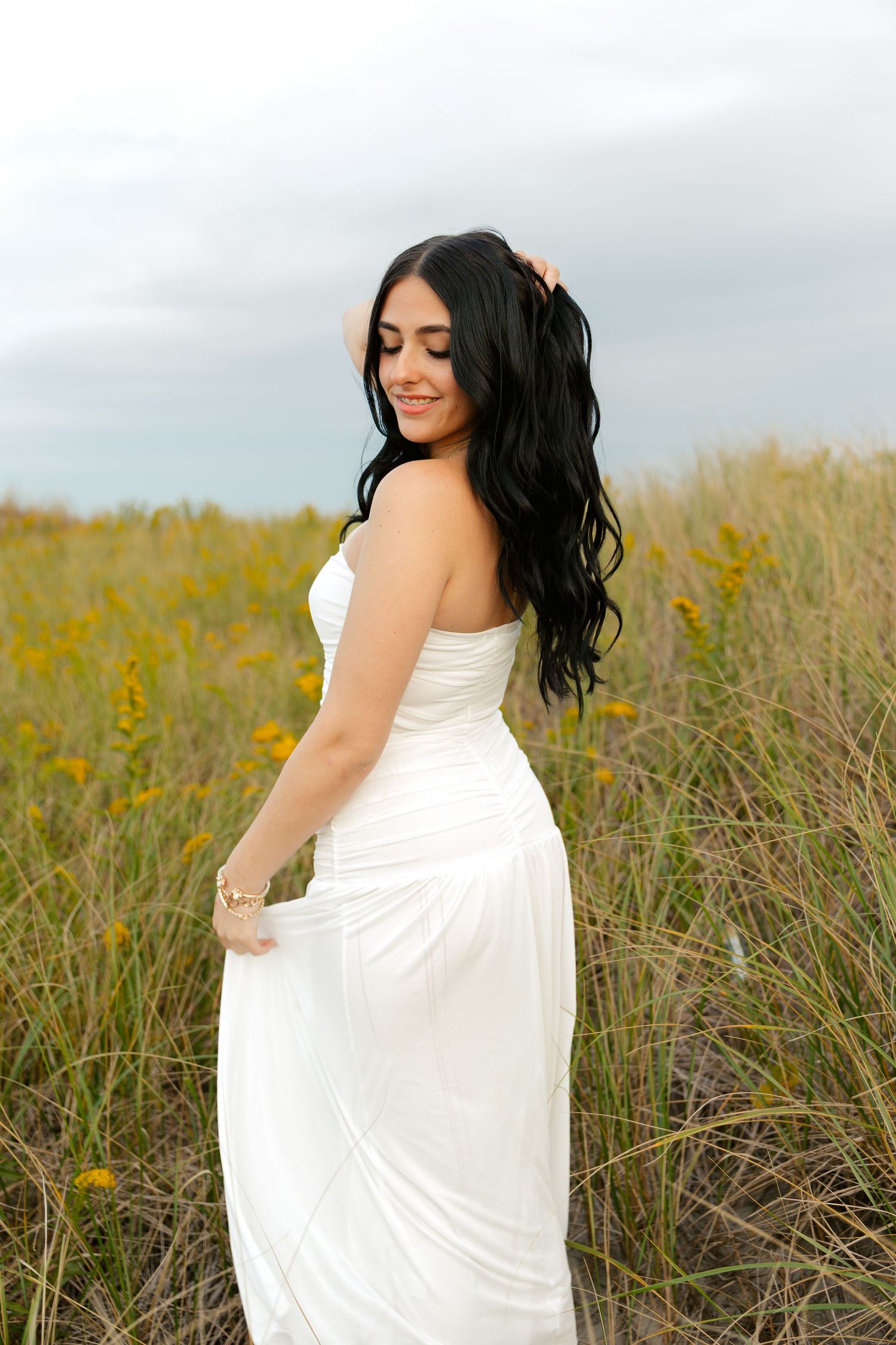 A woman in a white strapless gown poses in a field of tall grass and yellow wildflowers under a cloudy sky.