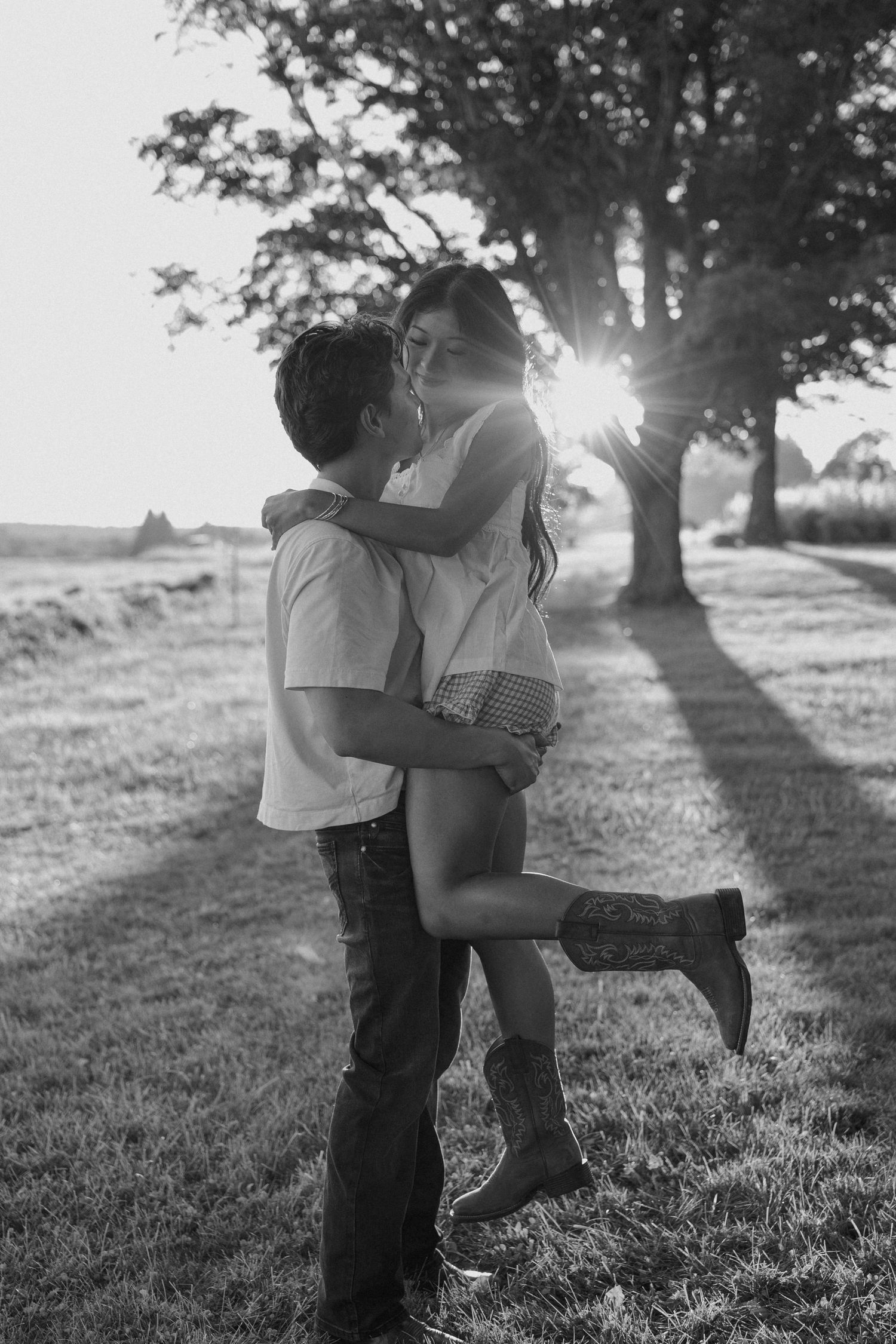 Black and white photo of a couple embracing outdoors at sunset, woman lifted off ground wearing cowboy boots.