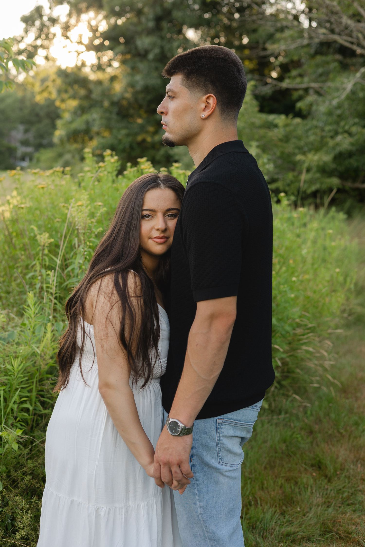 A young couple holds hands in a lush green field, woman in white dress, man in black shirt and jeans.