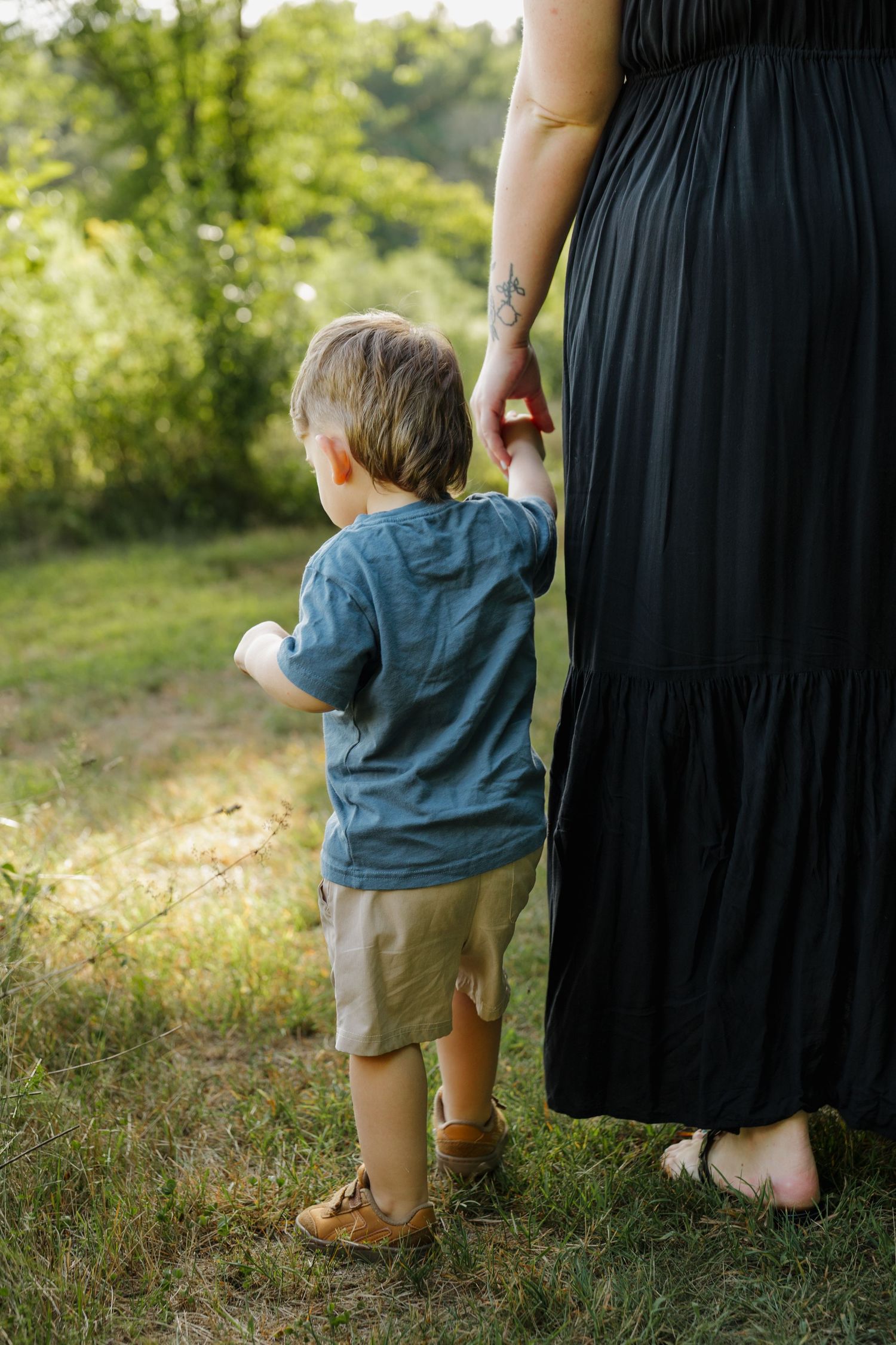 A young toddler holds his mother's hand while walking outdoors in a sunny green park, she wears a black maxi dress.