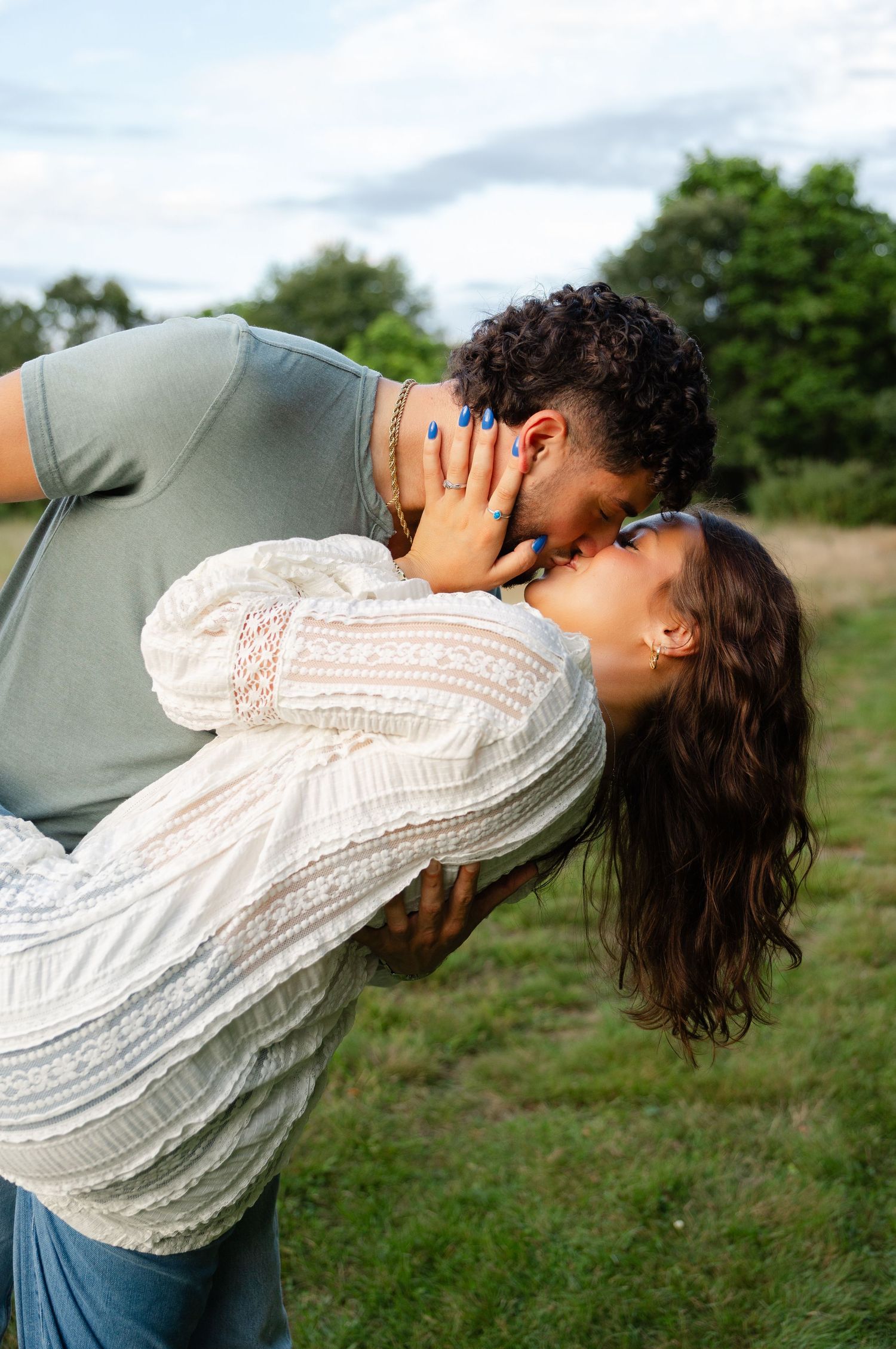 A couple shares a romantic kiss outdoors, with the woman in a white lace blouse leaning back in a green field.