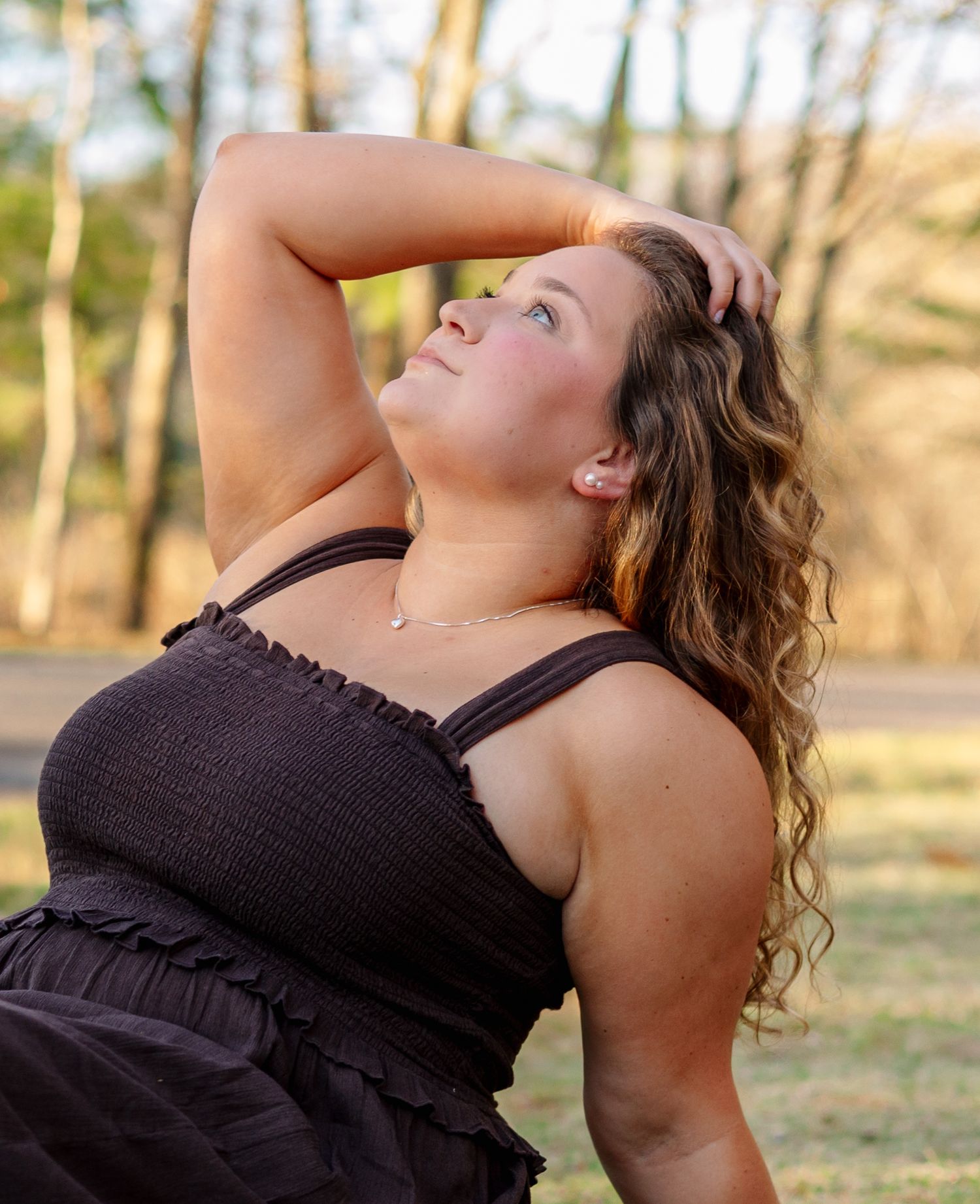 Young woman in black smocked sundress posing outdoors with hand behind head, looking up in warm sunset light.