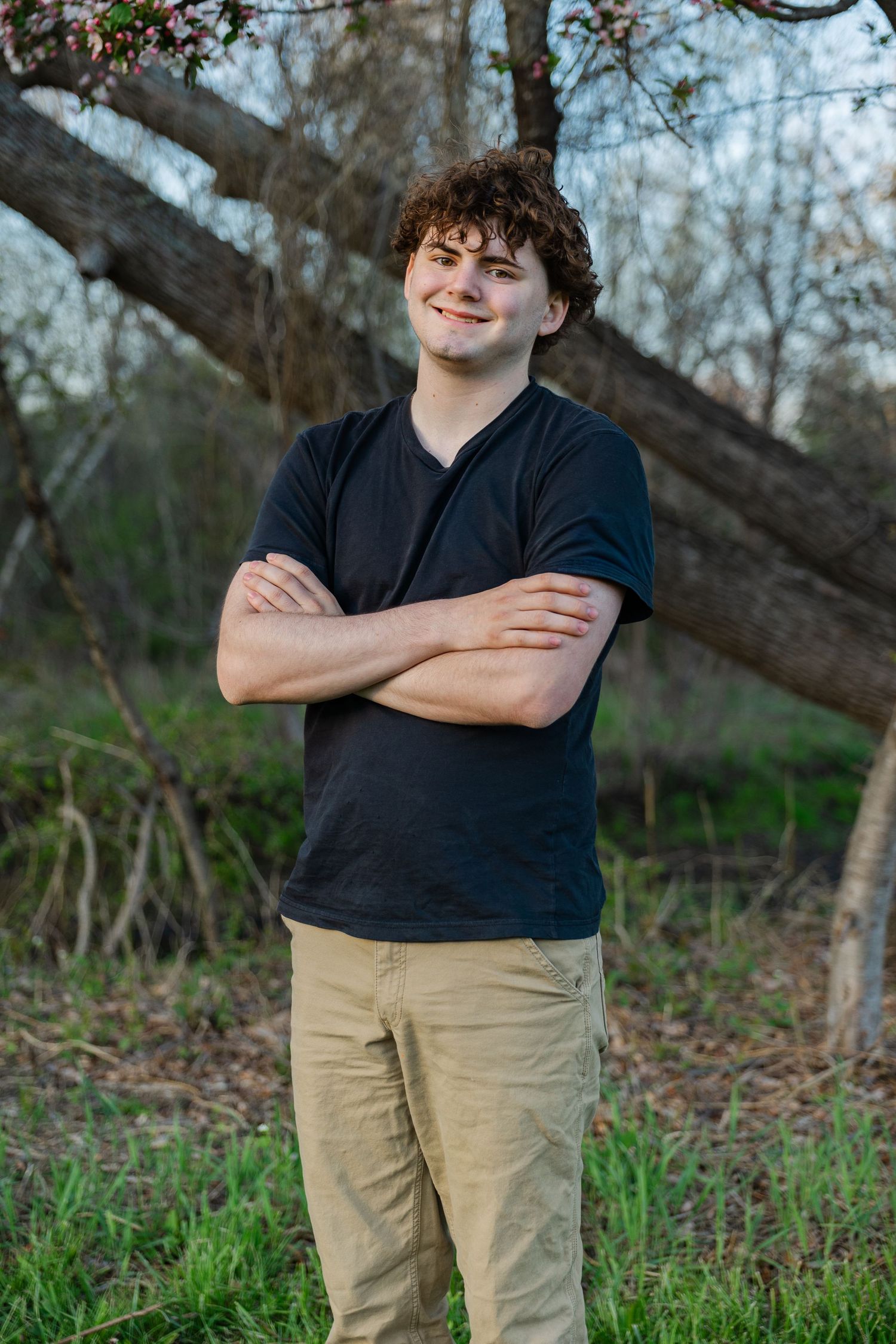 Smiling young man with curly hair standing arms crossed by tree, wearing black v-neck tee and khaki pants.
