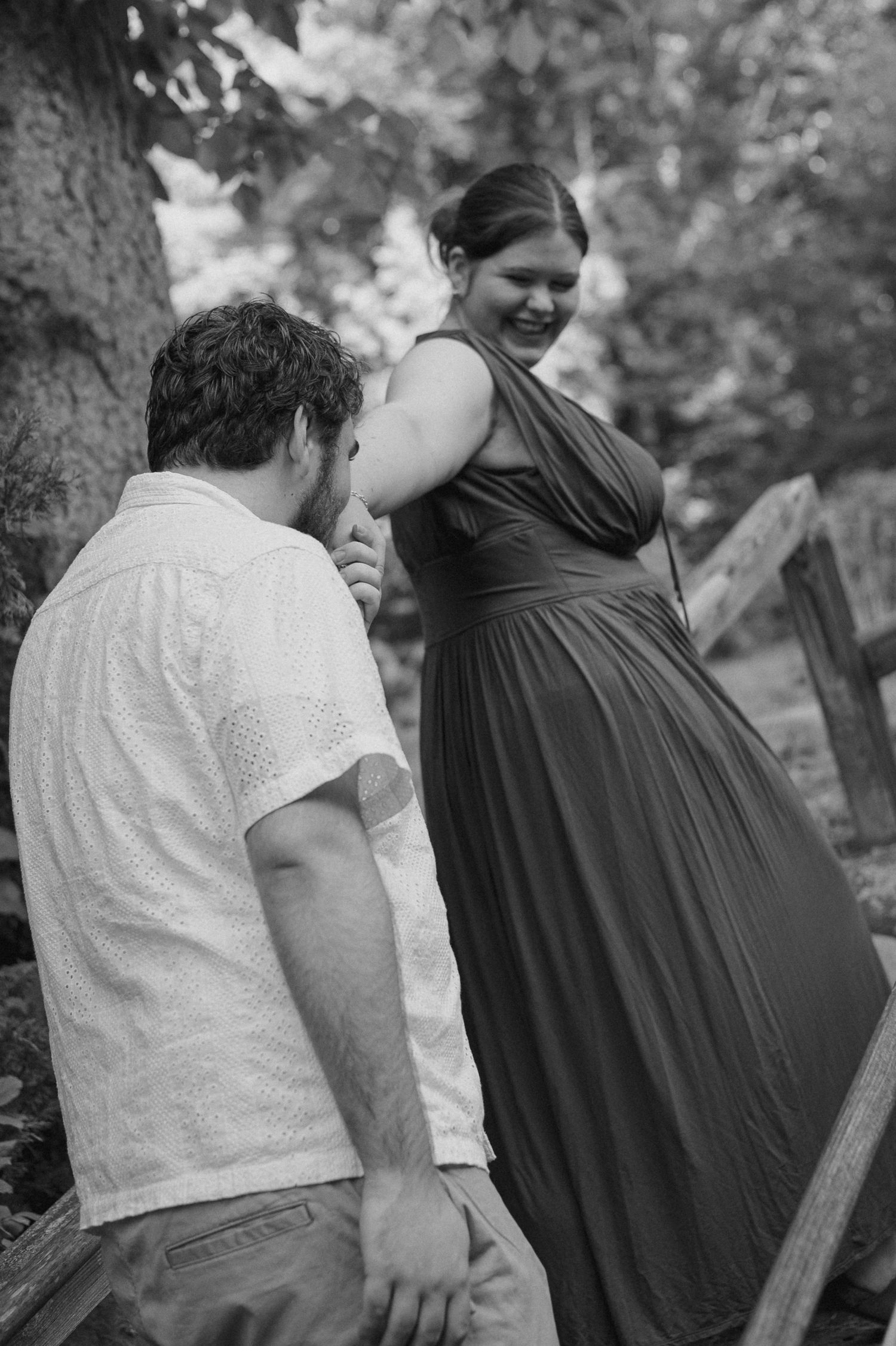 Black and white photo of a couple sharing a tender moment outdoors, woman in long dress smiling at man in white shirt.