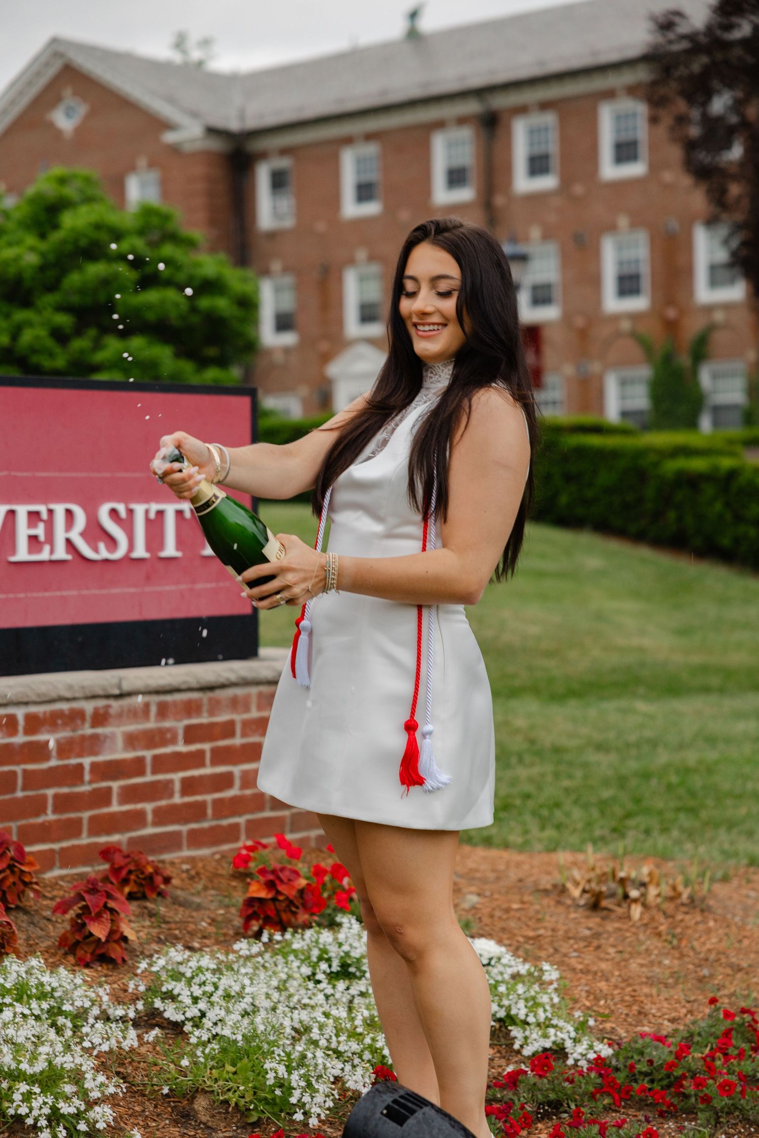 Young woman in white dress celebrating graduation by popping champagne bottle in front of university sign.