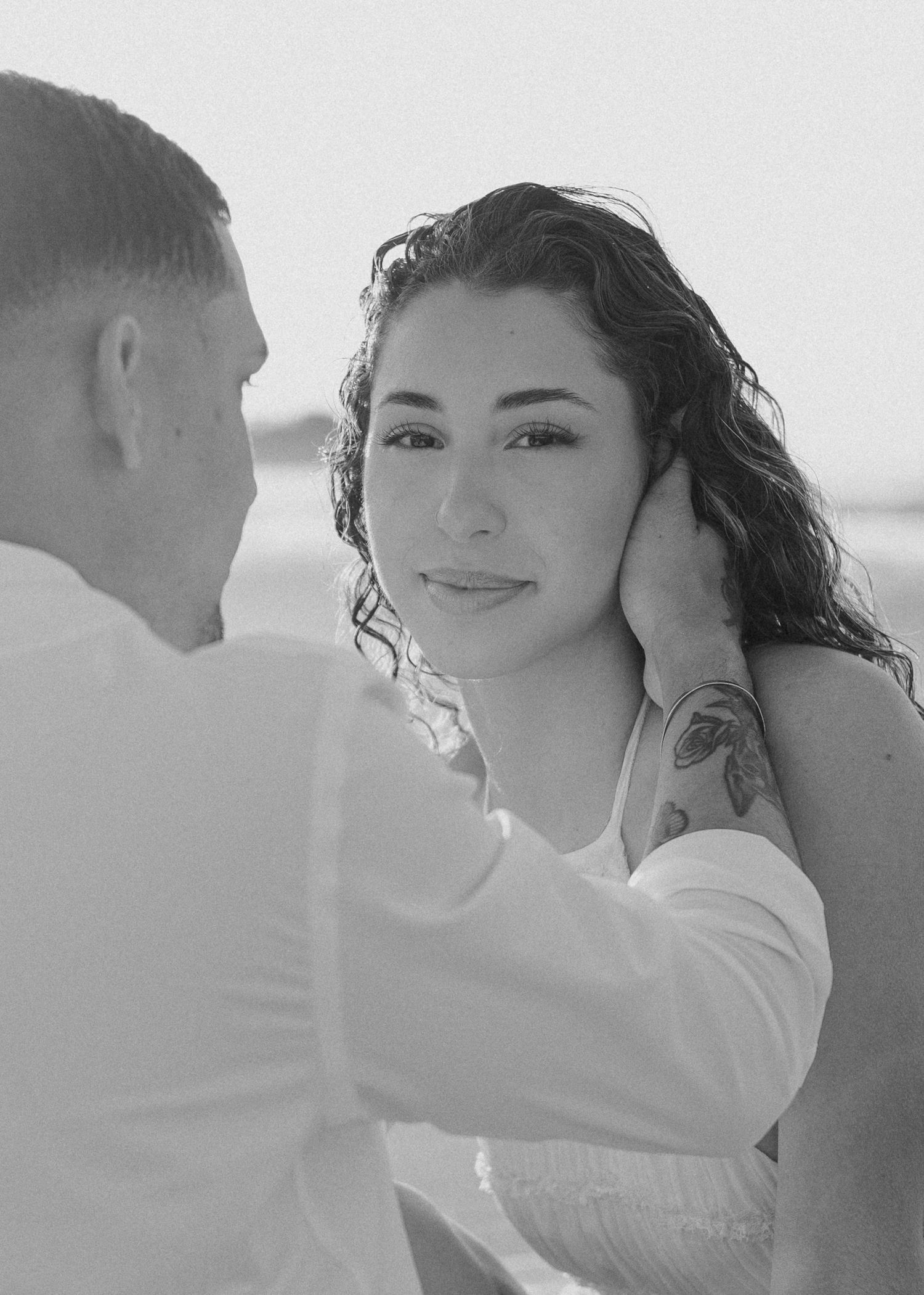 Black and white romantic beach photo of man gently holding womans face with curly hair and shoulder tattoo.