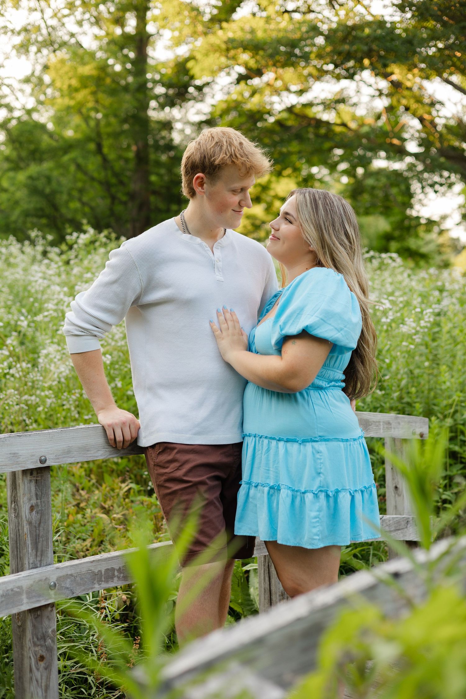 A young couple smiles at each other on a wooden bridge surrounded by lush greenery and wildflowers.