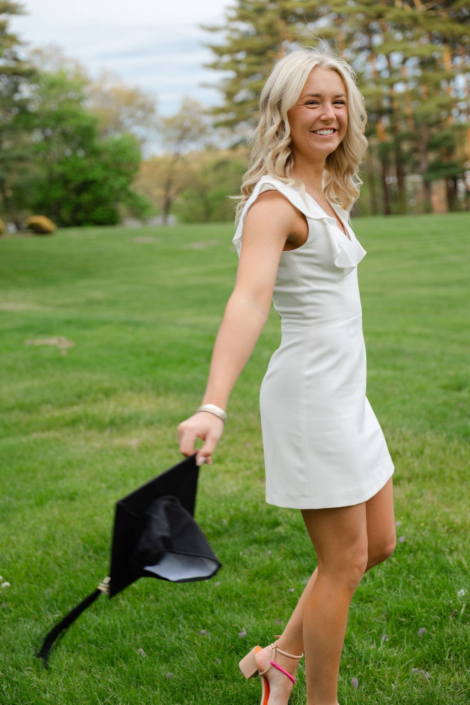 Smiling blonde woman in white dress holding graduation cap on green lawn.