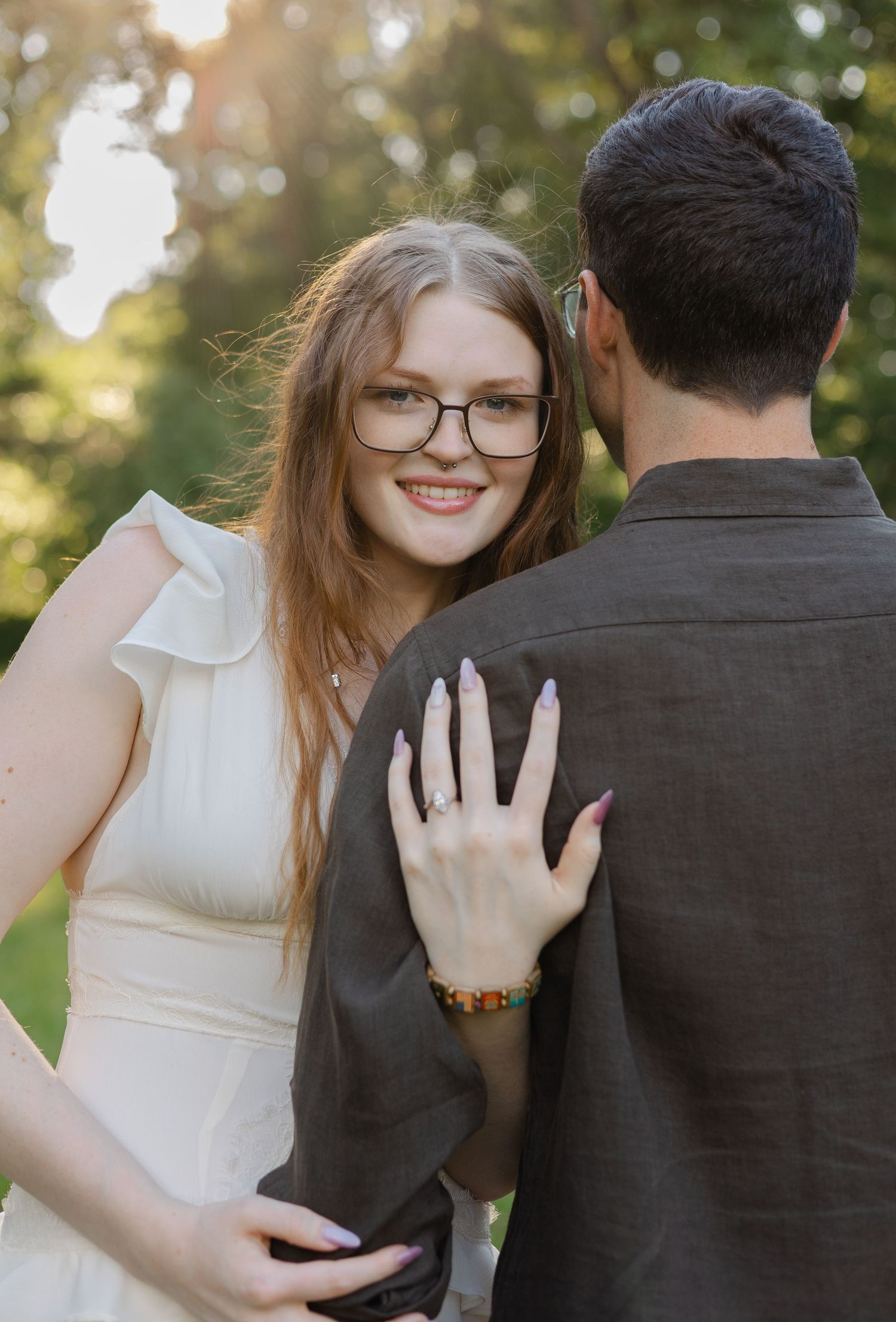 Smiling young woman in white dress and glasses rests hand on mans shoulder outdoors in sunlit park setting.