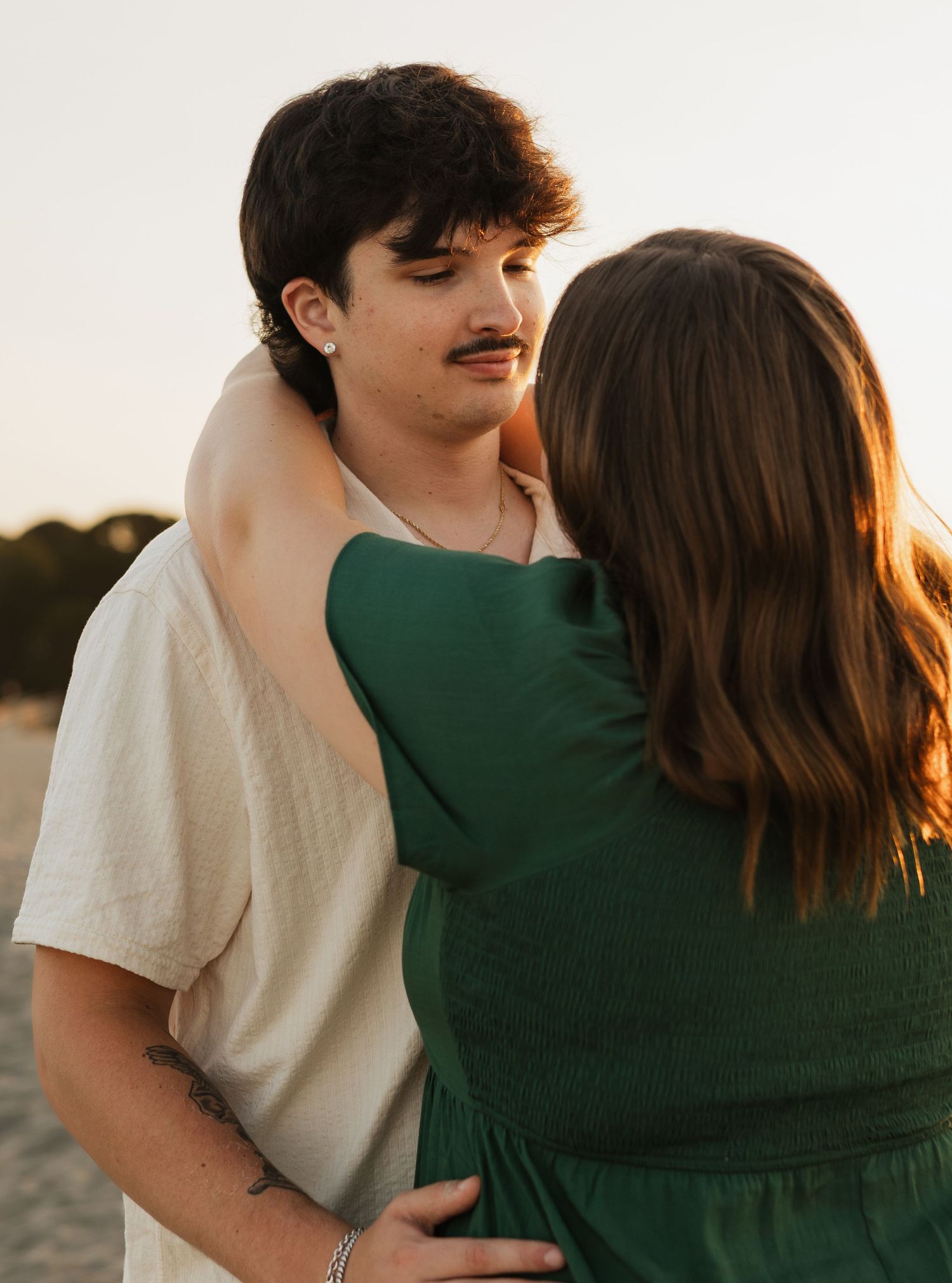 A young couple embracing at sunset, man in white shirt with mustache, woman in green dress, golden hour beach setting.