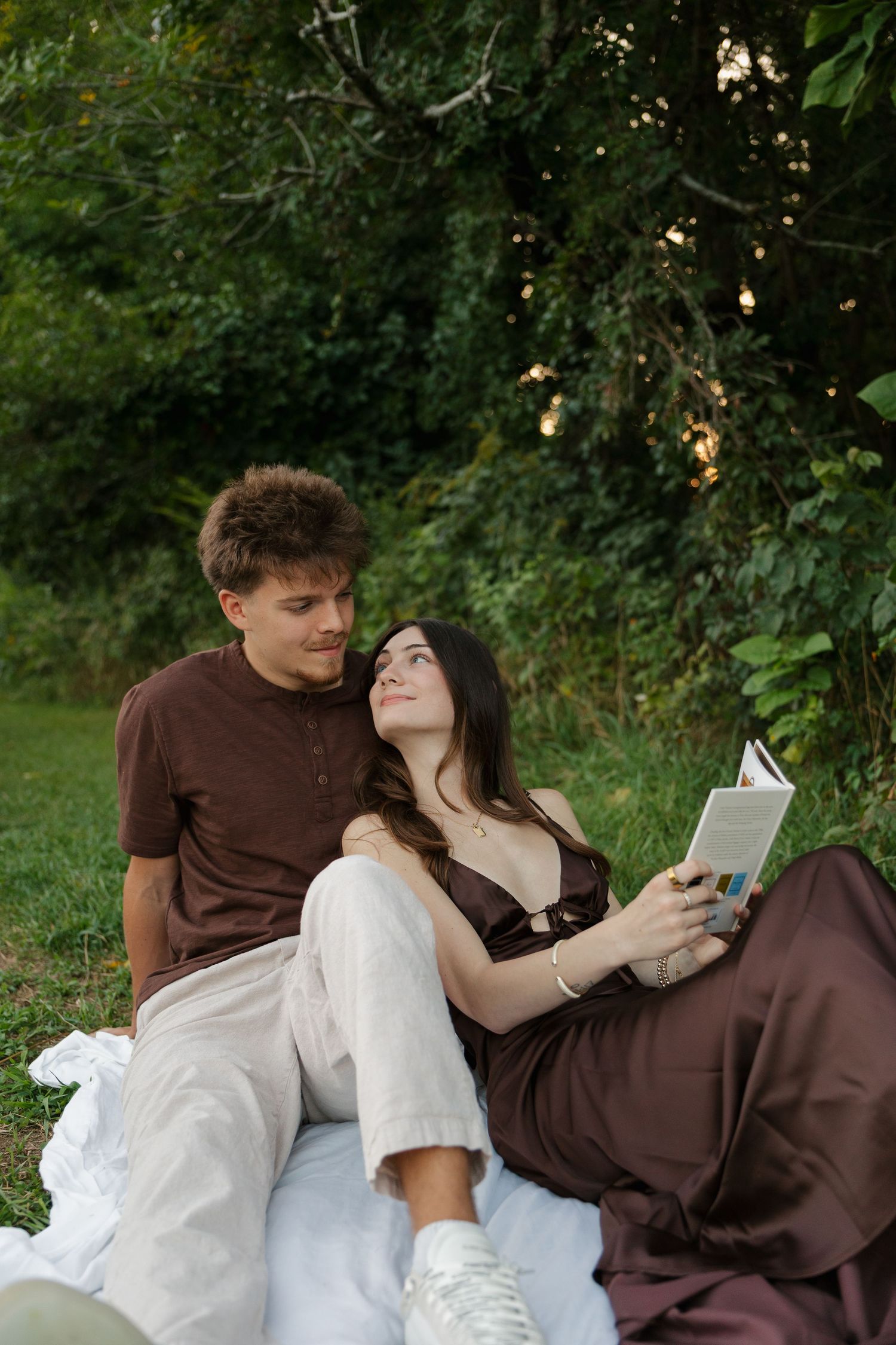 Young couple relaxing on a blanket outdoors, woman reading a book while leaning against her partner in nature.