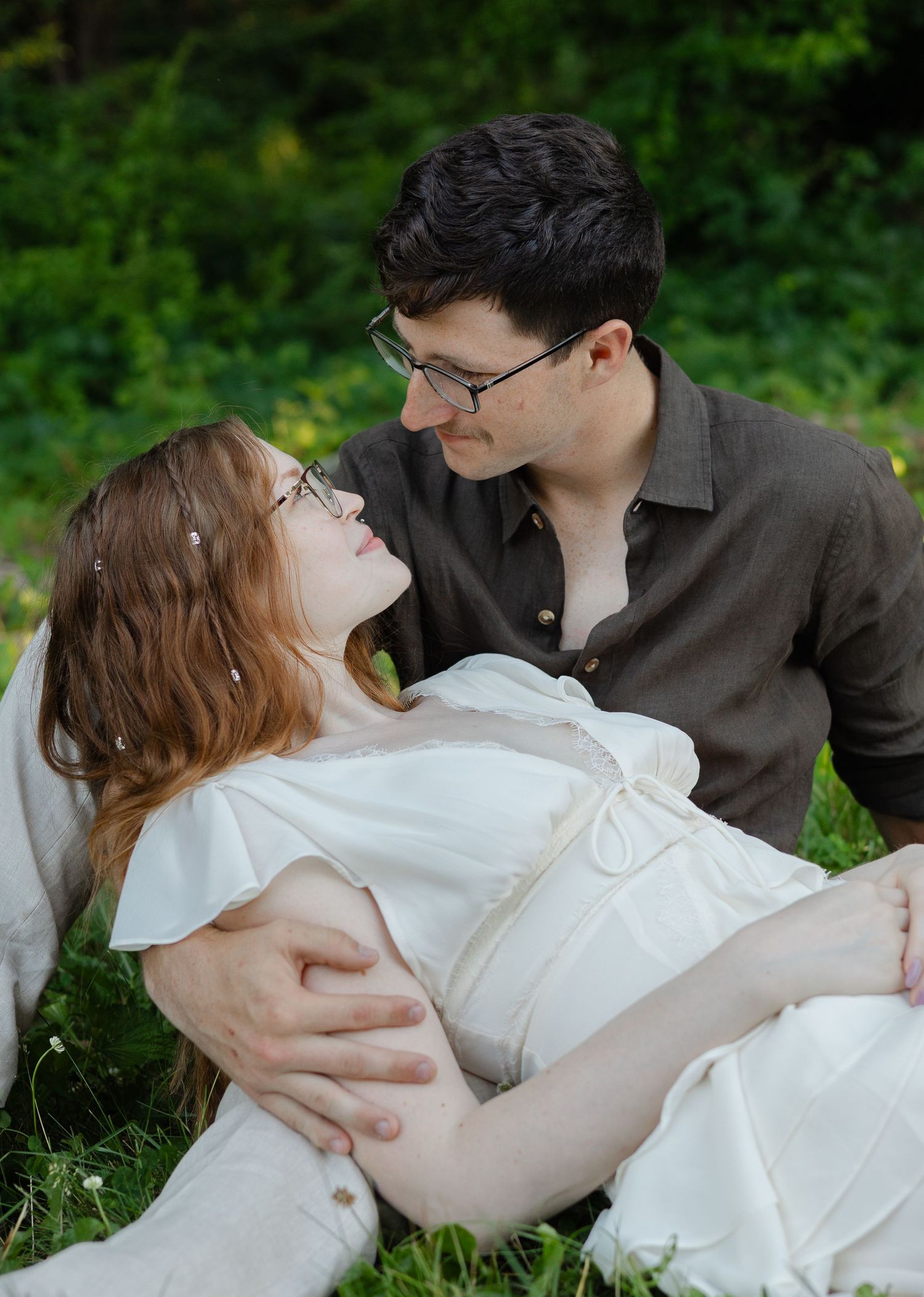 Couple sharing a tender moment on grass, pregnant woman in white dress lying back while partner gazes lovingly at her.