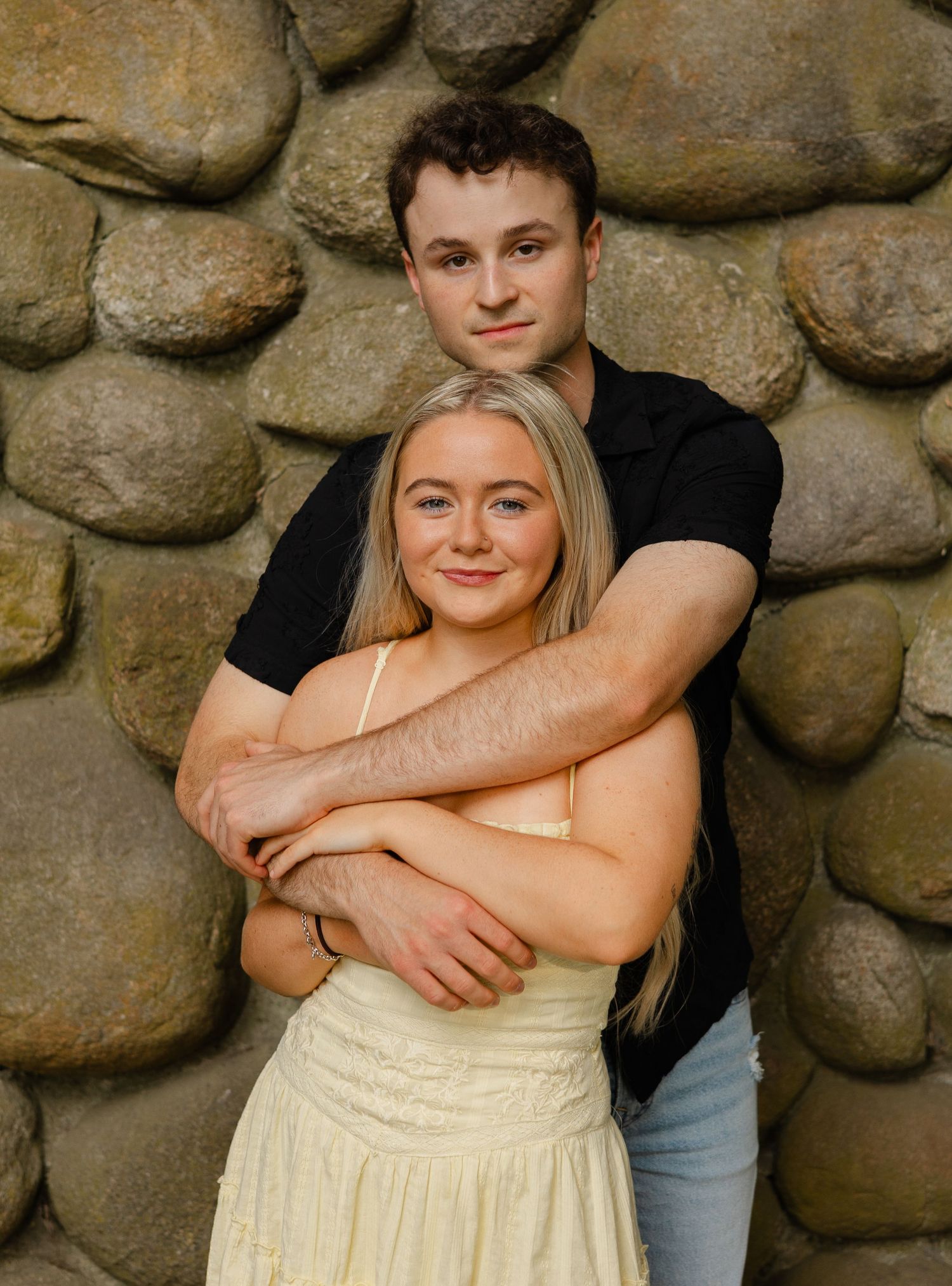 A young couple embracing in front of a stone wall, woman in yellow dress with long blonde hair.