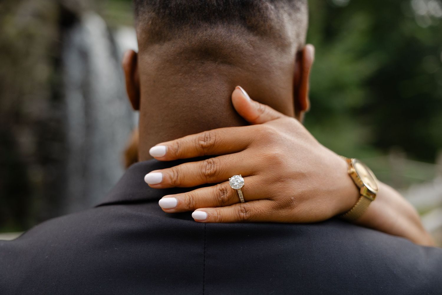 Woman with white manicure and diamond engagement ring embraces man outdoors near waterfall.